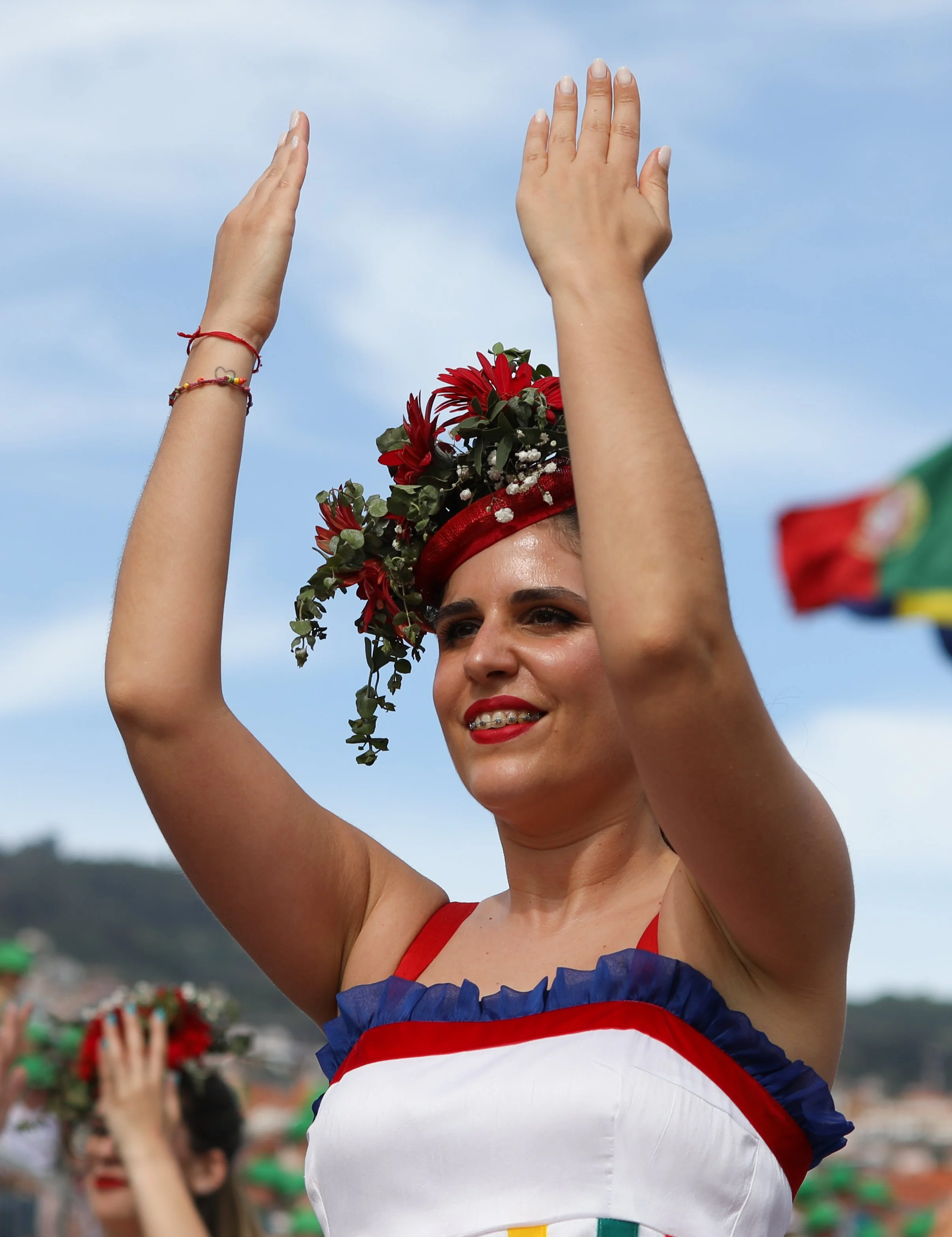 A woman dressed in traditional Portuguese attire, wearing a colorful dress with red, blue, and yellow accents, and a floral crown with red flowers and greenery, raising her arms in a celebration during a festival.