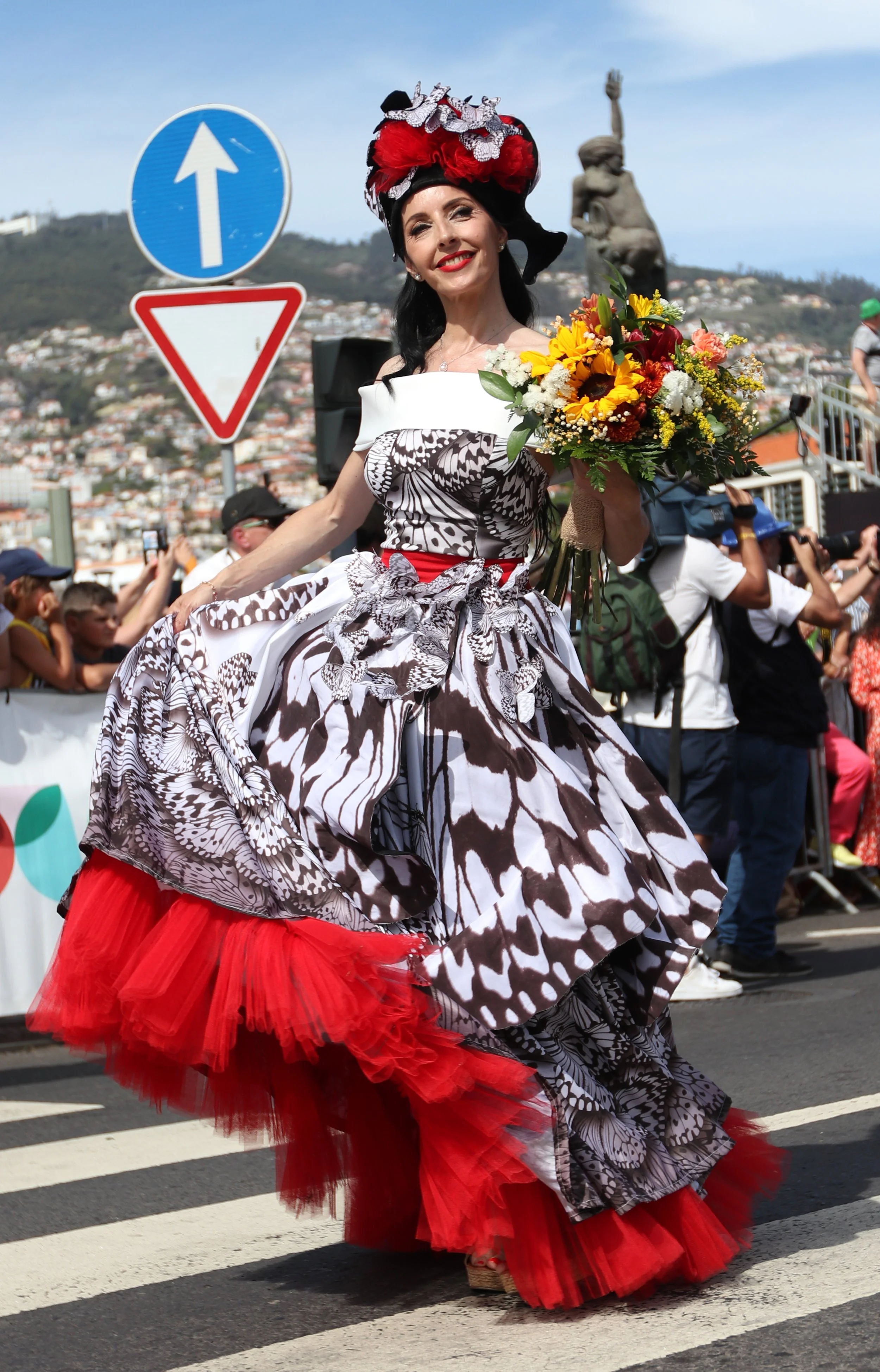 Woman in a vibrant, patterned dress and red petticoat holding a bouquet of flowers, walking on a street during a parade or festival, with an audience and photographers in the background, and a scenic hillside with buildings and statue behind.