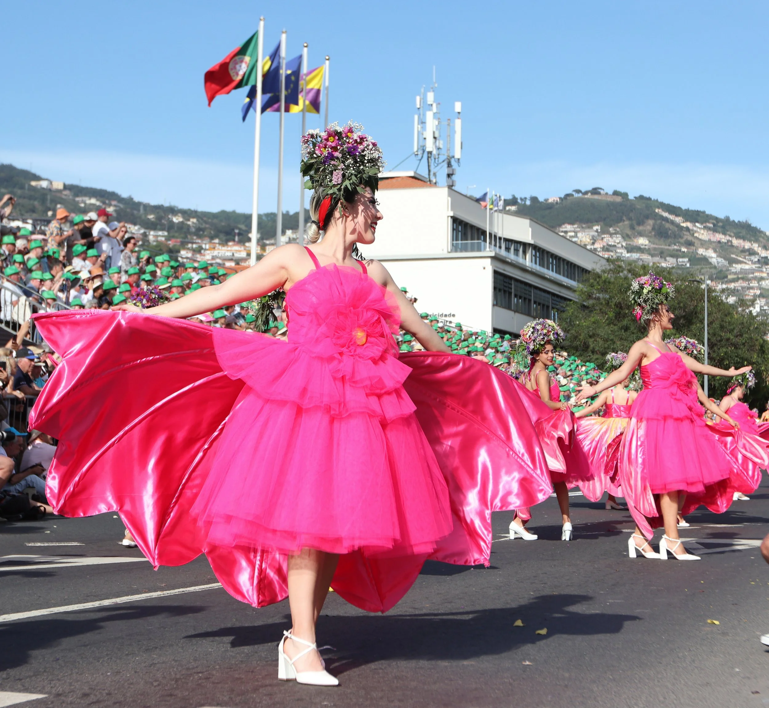 Women in bright pink dresses with floral headpieces performing a parade on a city street.