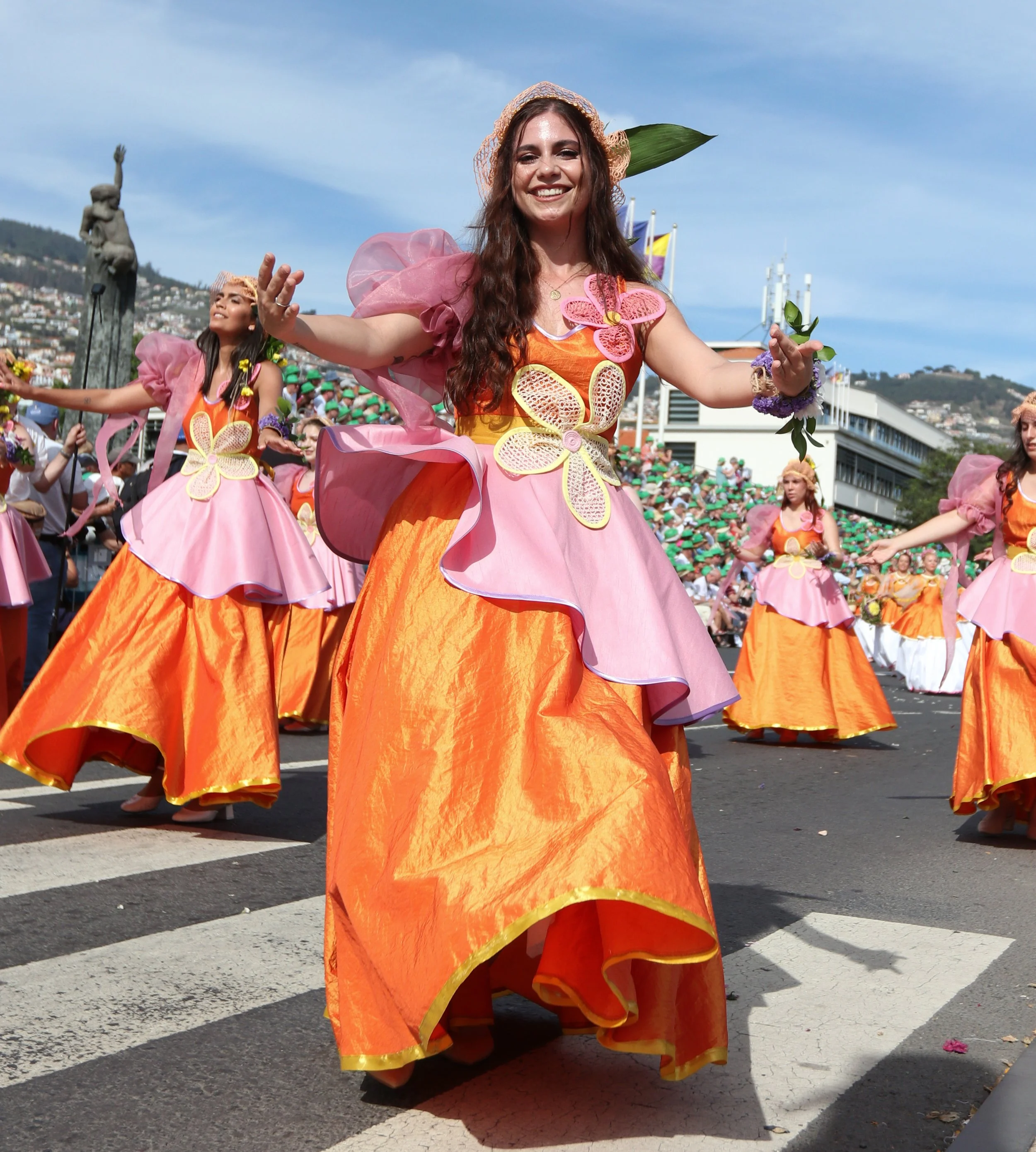 A woman in a colorful orange and pink dress dancing in a parade, surrounded by other women in similar costumes, with a crowd of spectators and a statue in the background.