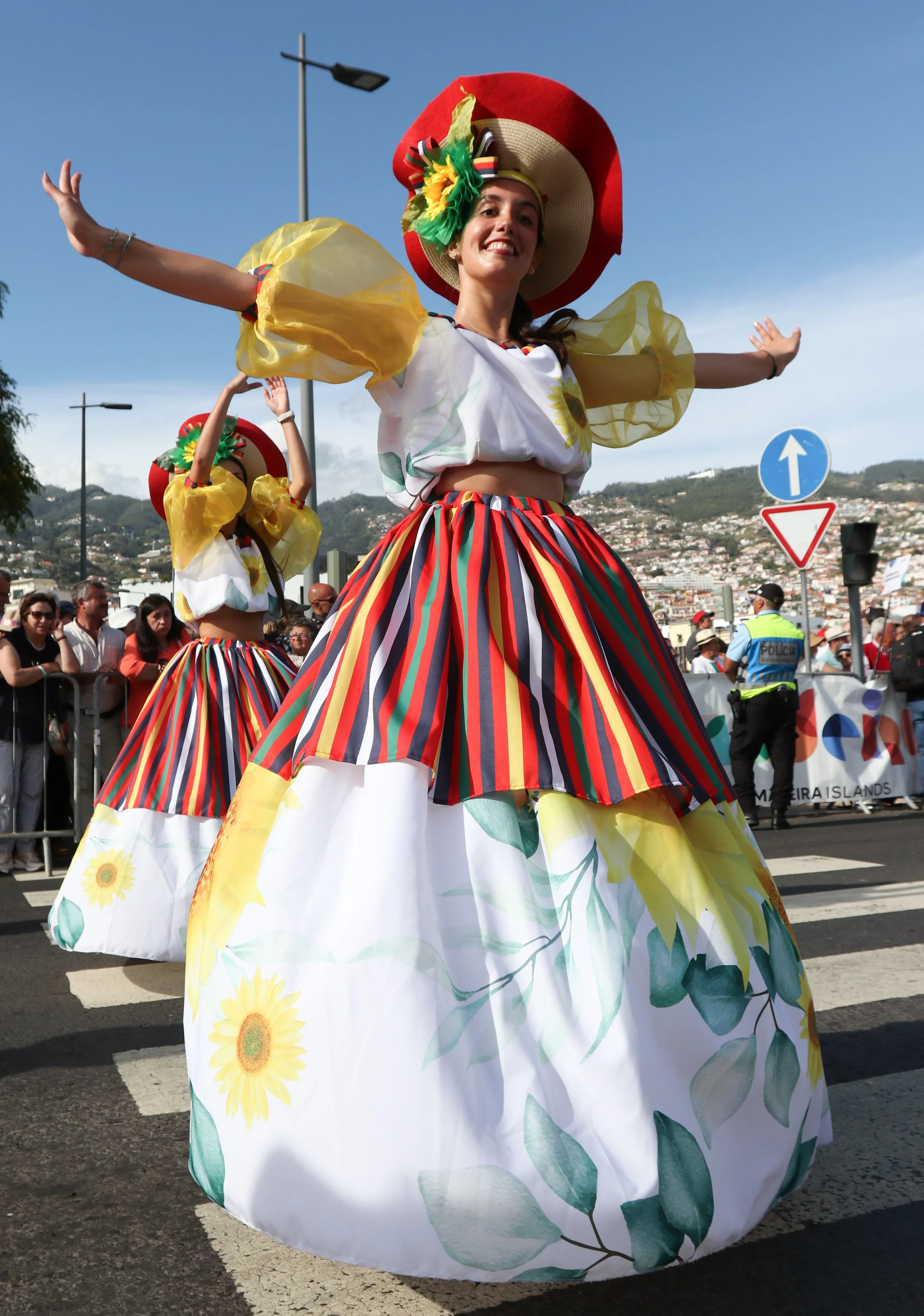 Women in traditional folk costumes dancing in a parade, wearing colorful striped skirts, floral tops, and large sun hats, with a crowd and police officers in the background.