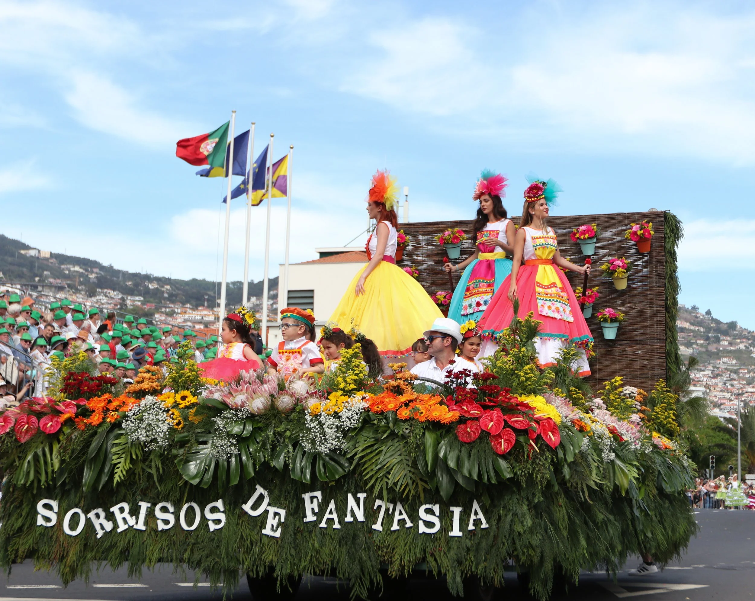 A colorful parade float decorated with flowers and traditional costumes, with women in vibrant dresses and feathered headpieces, titled 'Sorrisos de Fantasia' in Portuguese, with flags flying in the background.