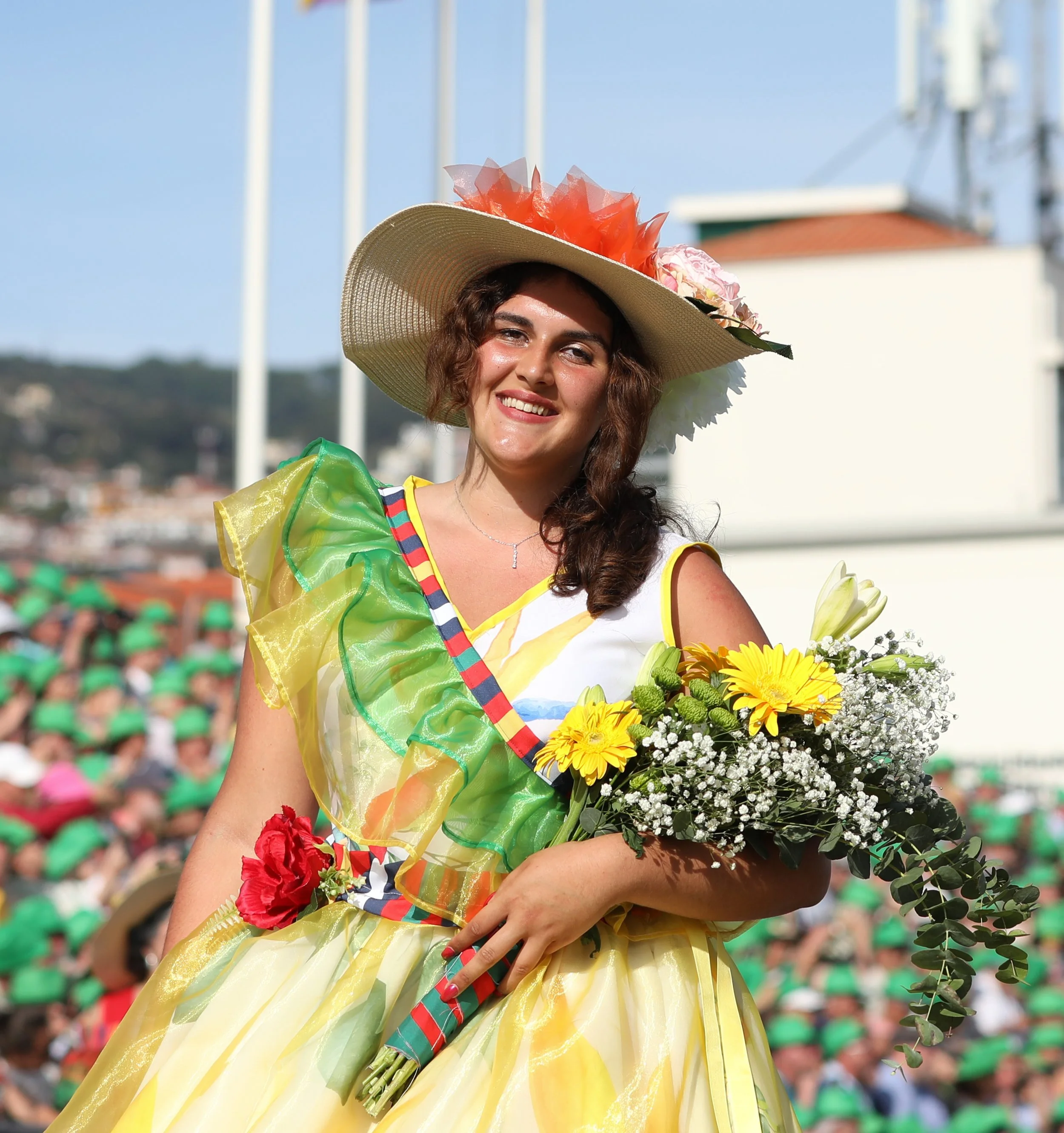 Woman in festive dress holding bouquet of flowers, smiling, wearing a large sun hat with floral decorations, during an outdoor celebration.