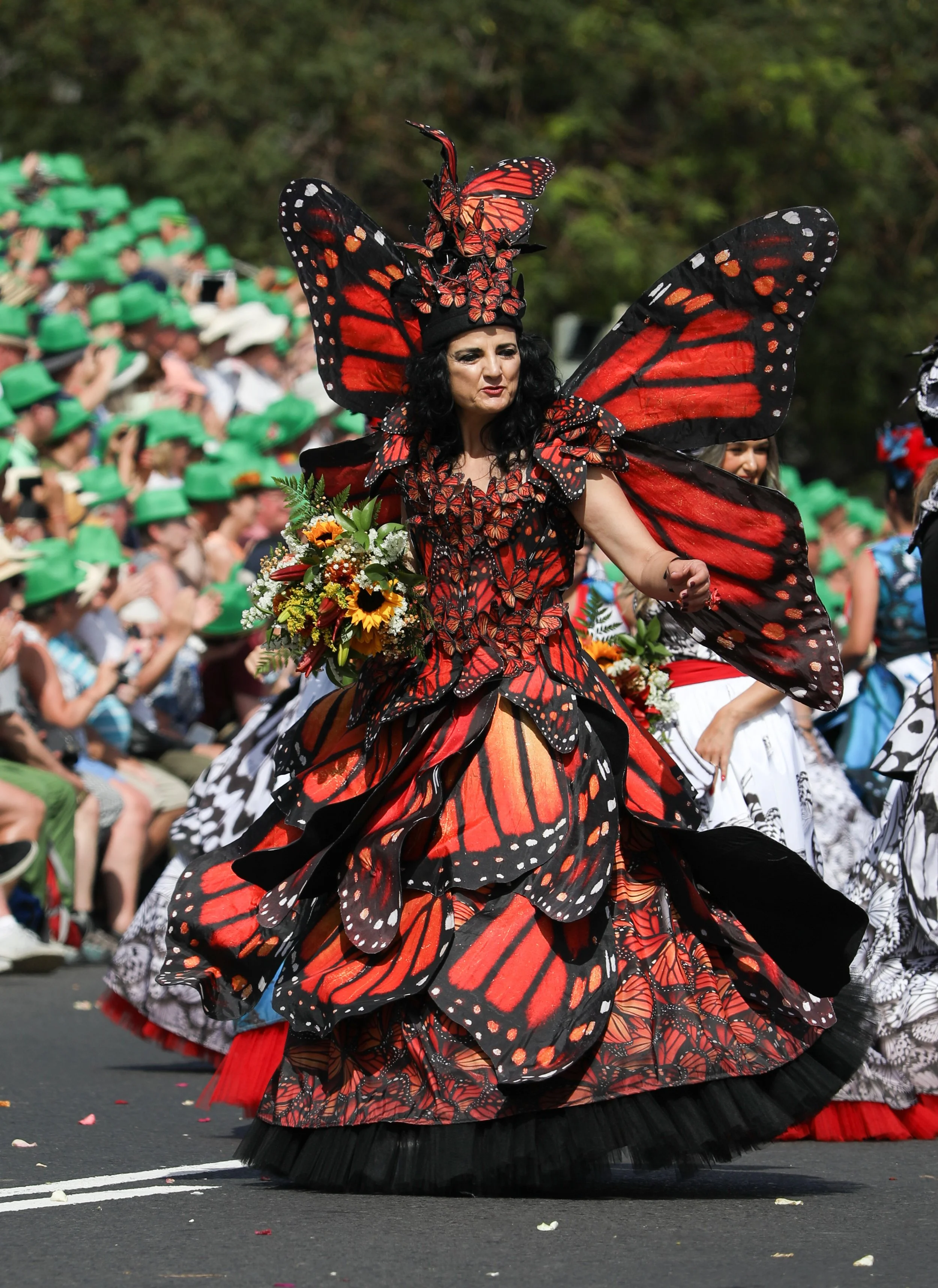 A woman in a butterfly costume with large red and black wings and dress, carrying a bouquet, participating in a parade with spectators wearing green hats in the background.