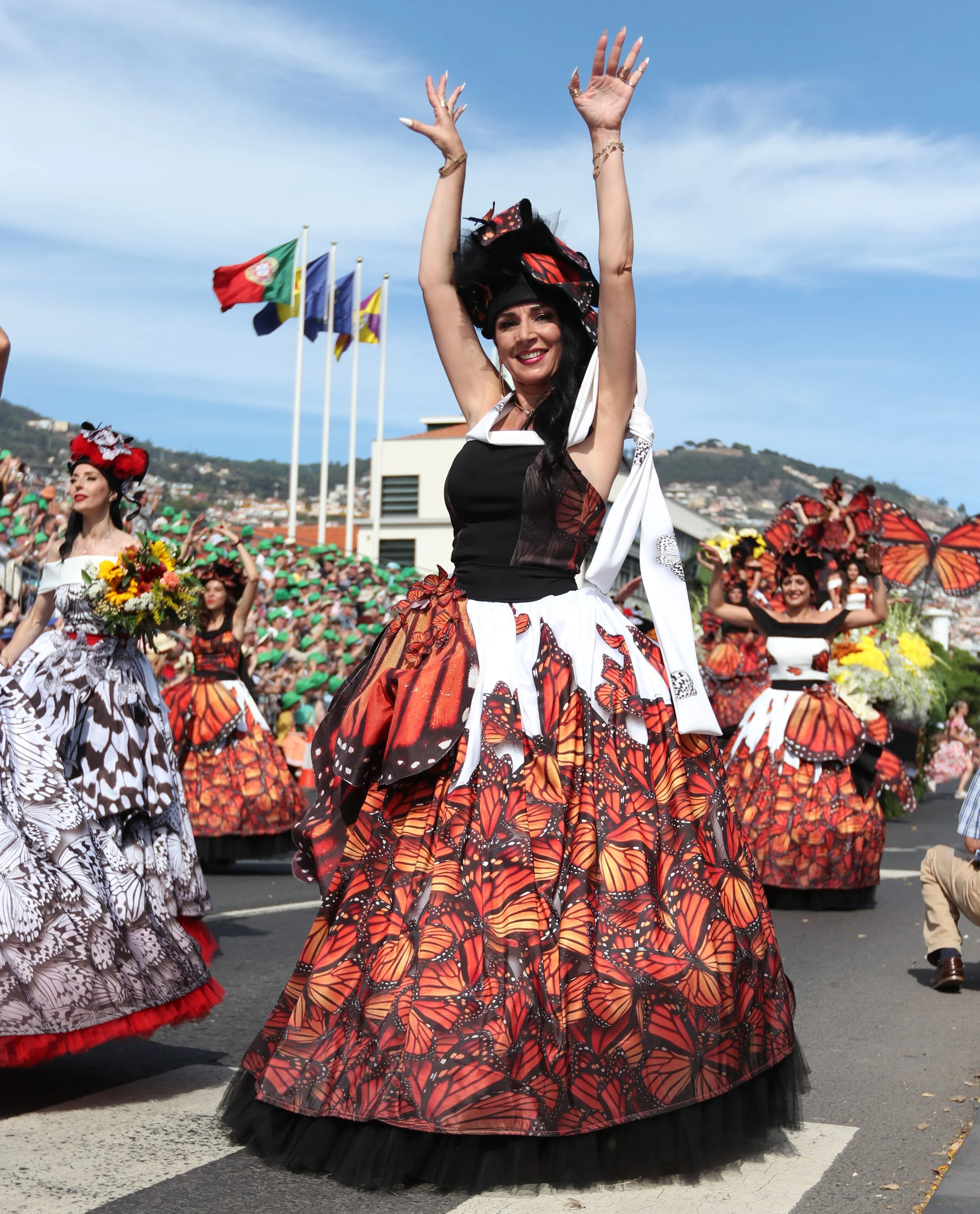 Women dressed in butterfly-themed costumes participating in a parade, with some holding flowers and butterfly wings, and flags flying in the background.