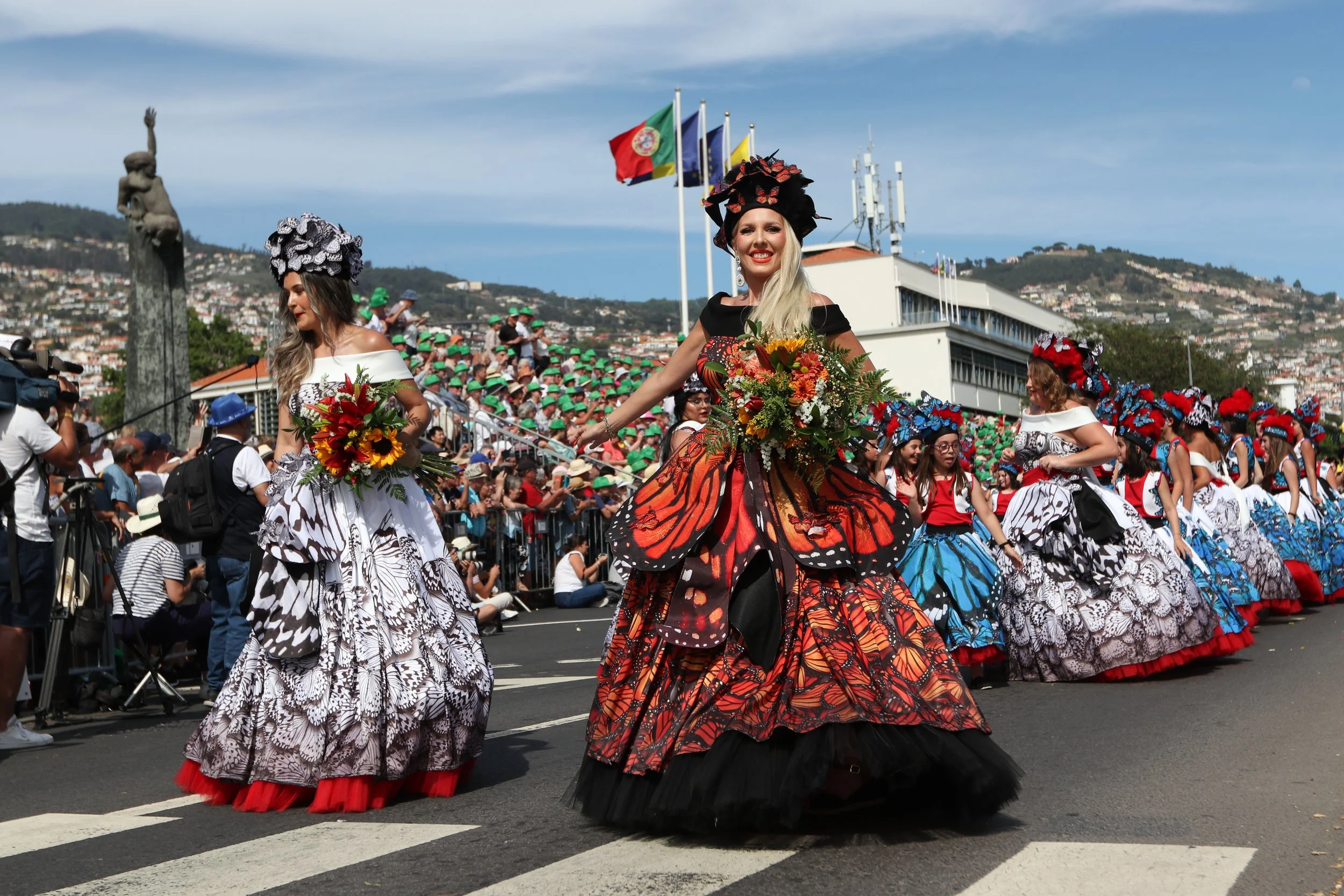 Women in colorful butterfly-themed dresses participating in a parade with onlookers and flags in the background.