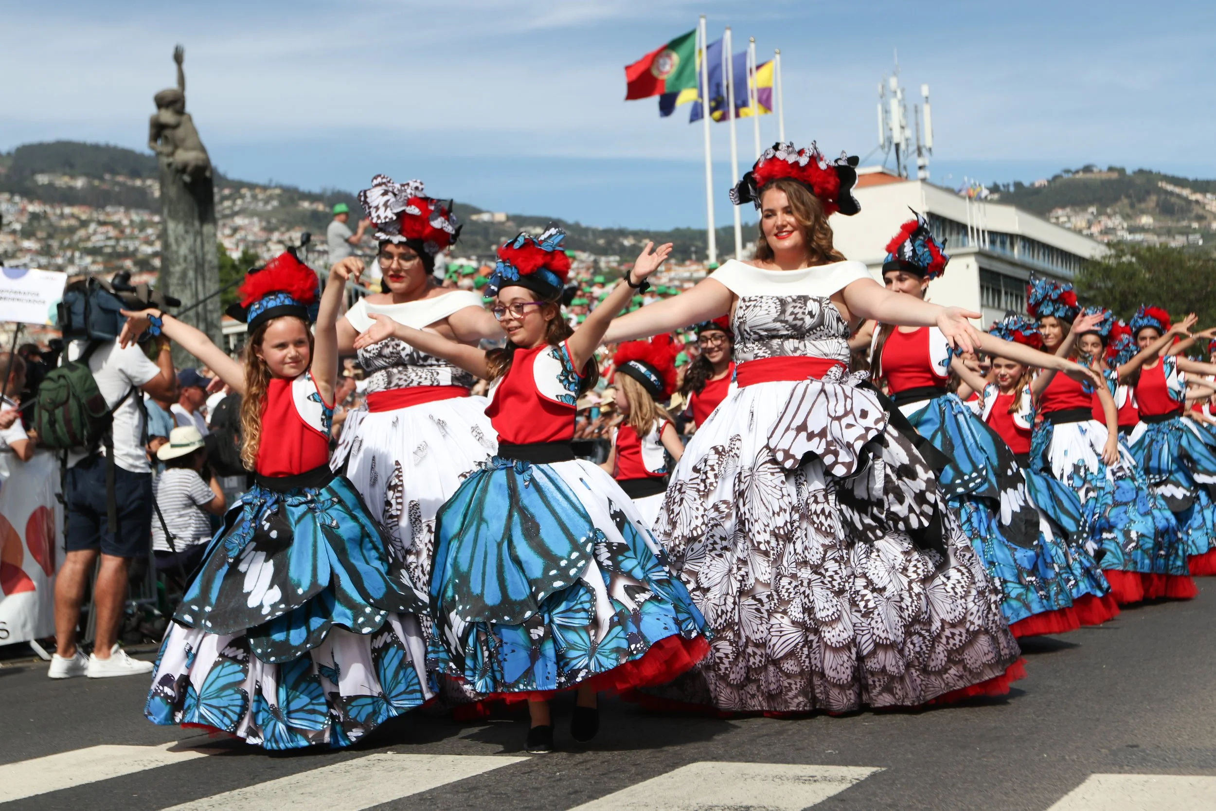 Women and girls in traditional butterfly-themed dresses and floral headpieces participating in a parade or festival, with a crowd of spectators and flags in the background.