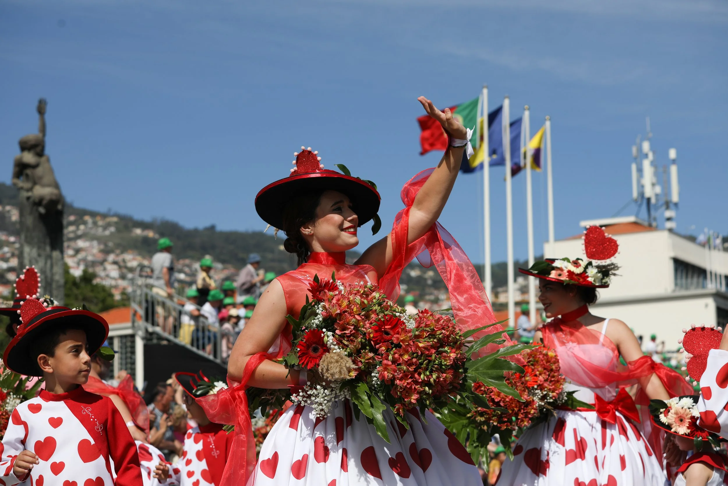 Women and children dressed in red and white costumes with heart patterns, participating in a parade with flowers, flags, and a crowd in the background.