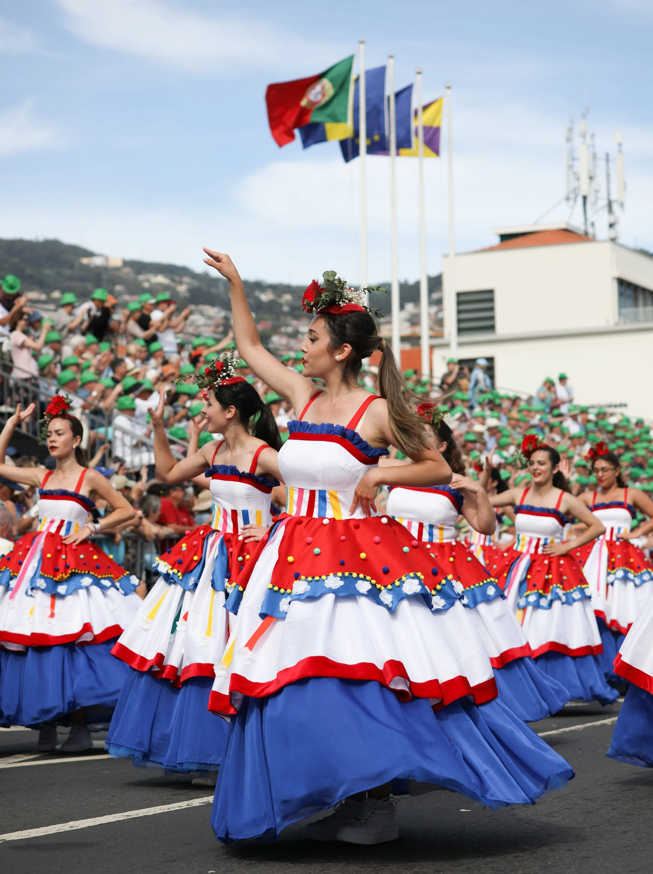 Women in colorful traditional dresses dancing in a parade with flags in the background.