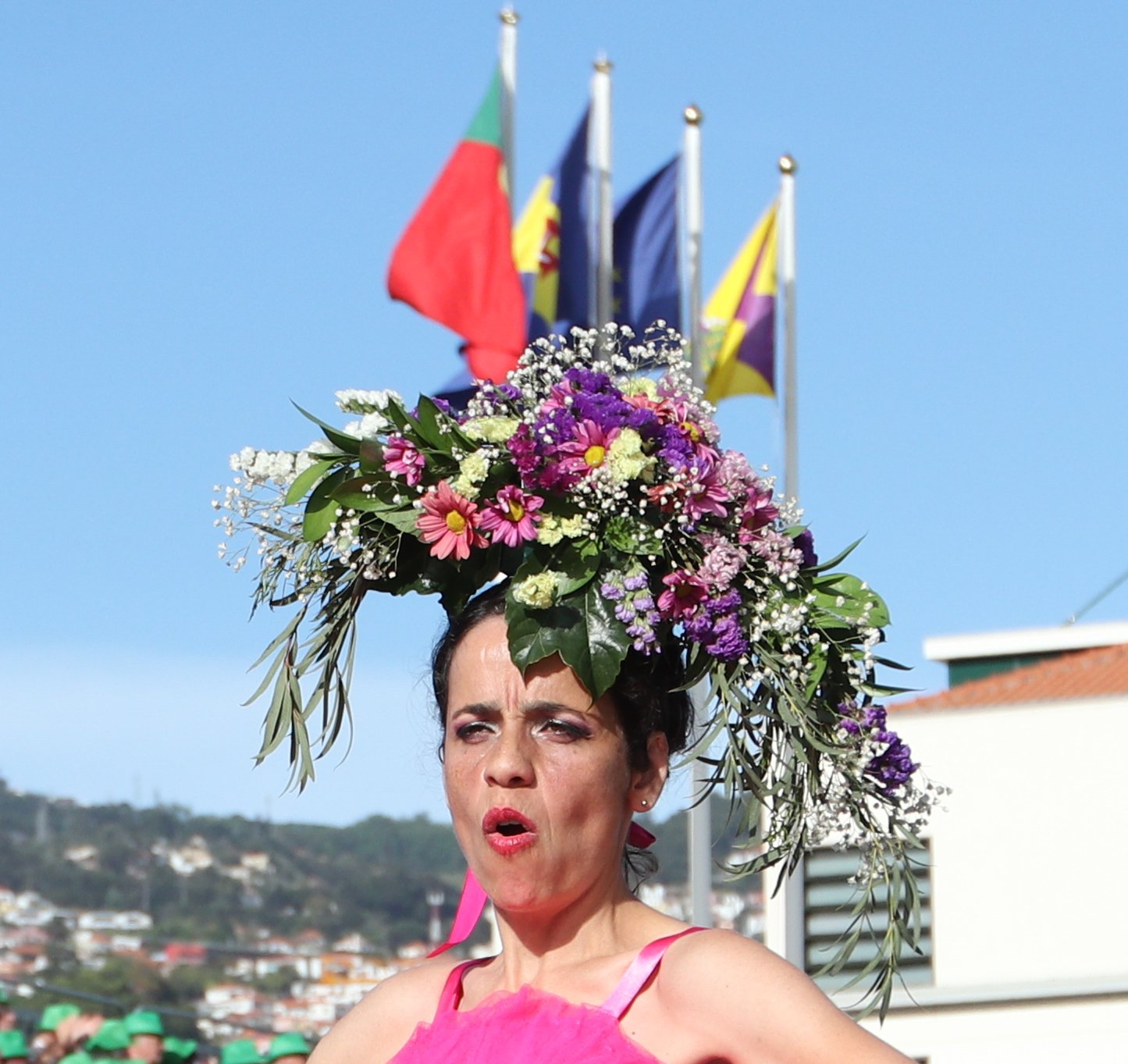 A woman wearing a large floral headpiece and a pink top, standing outdoors with flags in the background.