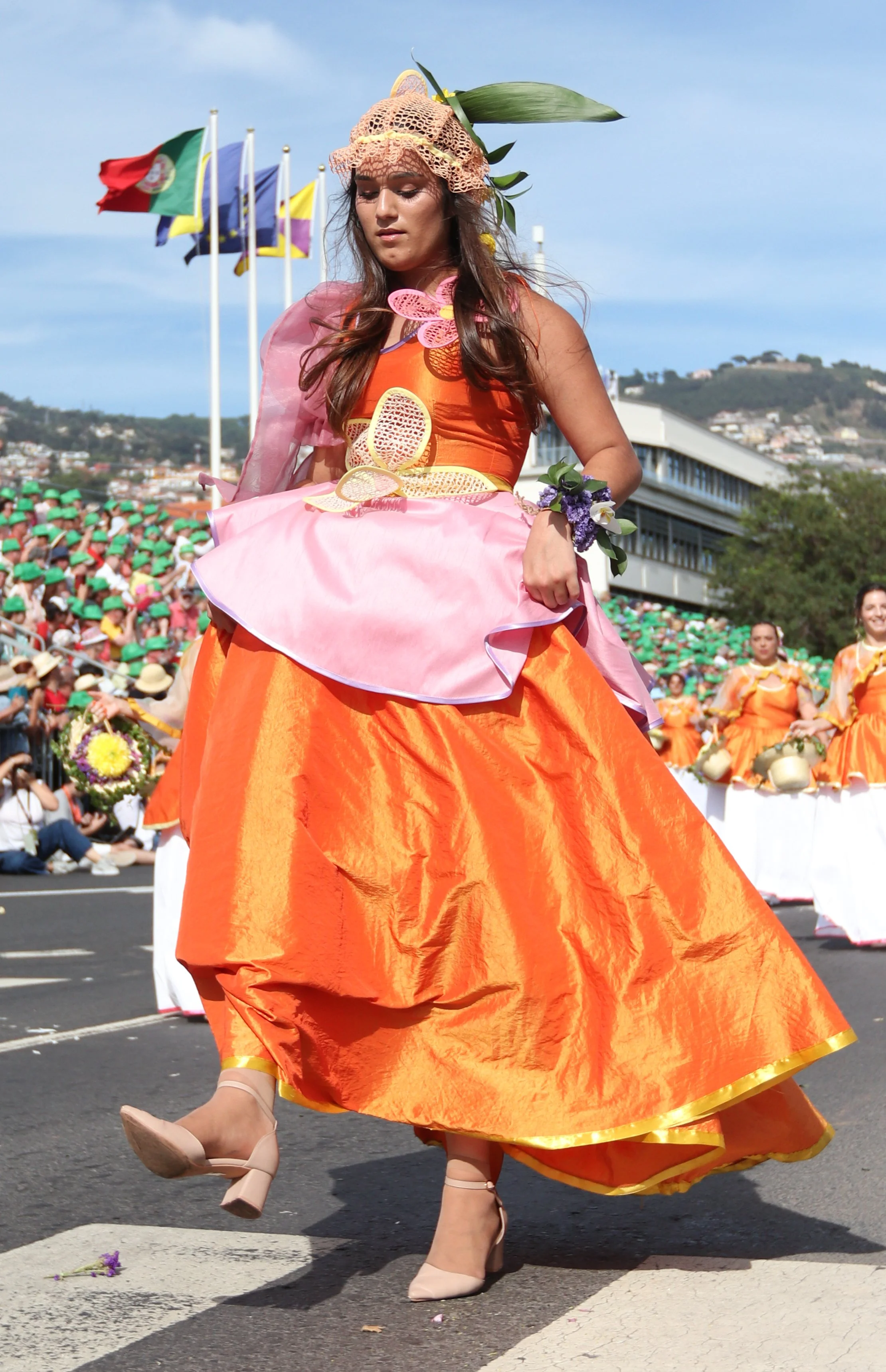 A woman in a colorful, traditional dress participating in a parade, with other women dressed similarly in the background and flags flying in the sky.