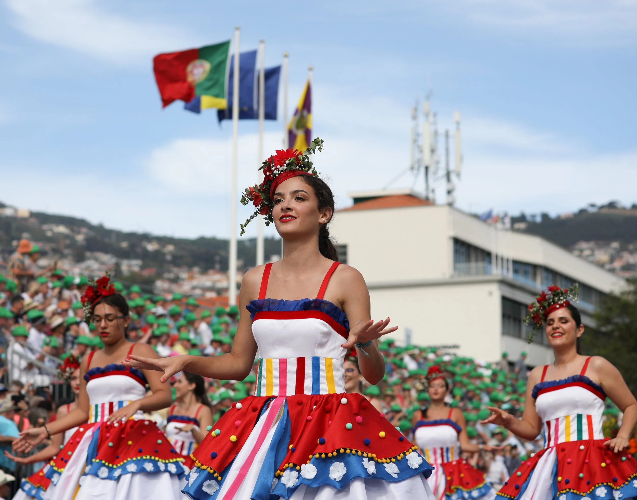 Women in colorful traditional dresses dancing in a parade with flags and a crowd in the background.