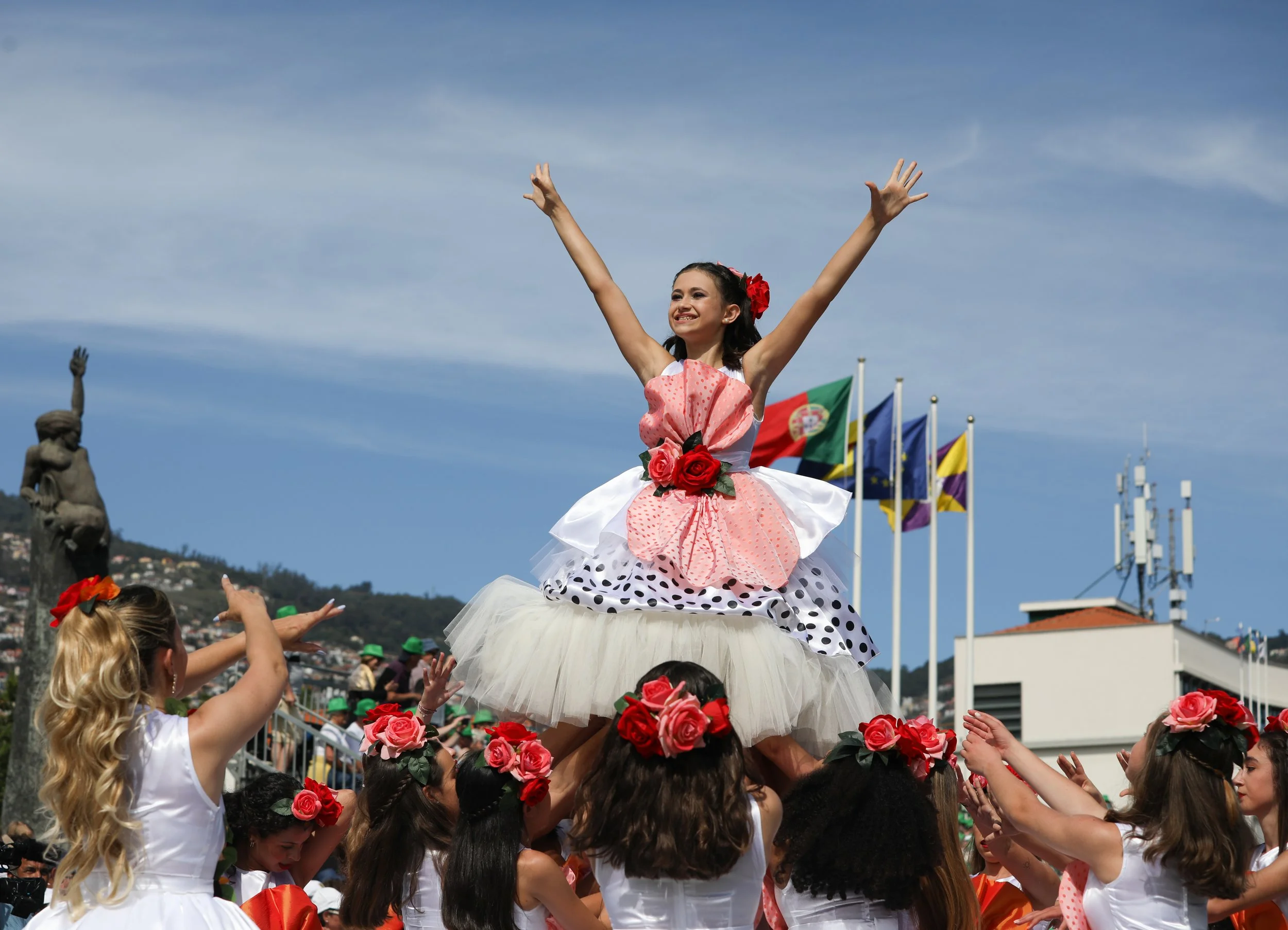 A young girl in a pink and white dress with red roses on the front and a polka dot petticoat is being lifted on the shoulders of others. She is smiling and raising her arms in celebration at an outdoor event with flags in the background.