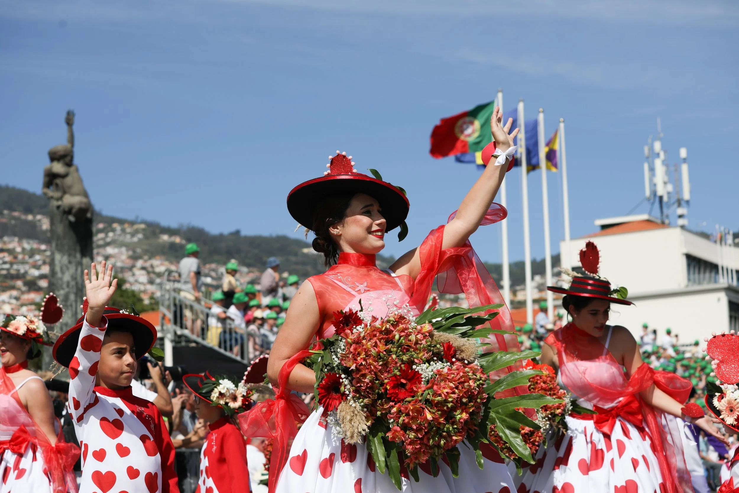 Women and children dressed in red and white with heart patterns, celebrating in a parade with flags, one woman holding a large bouquet of red and white flowers, and a woman waving.