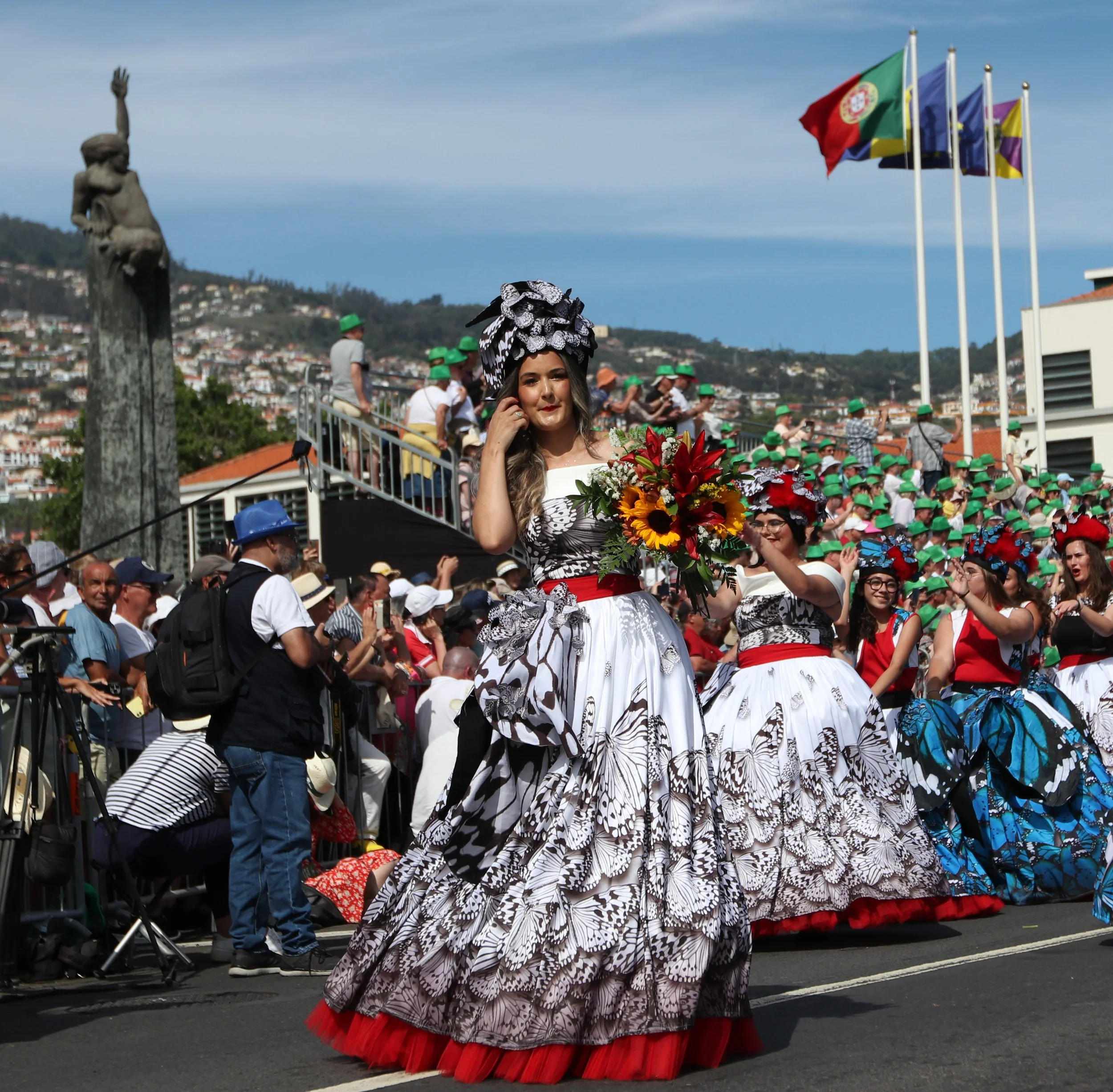 Women in butterfly-patterned traditional dresses and headpieces holding flowers participate in a parade on a city street, with a crowd and flags in the background.