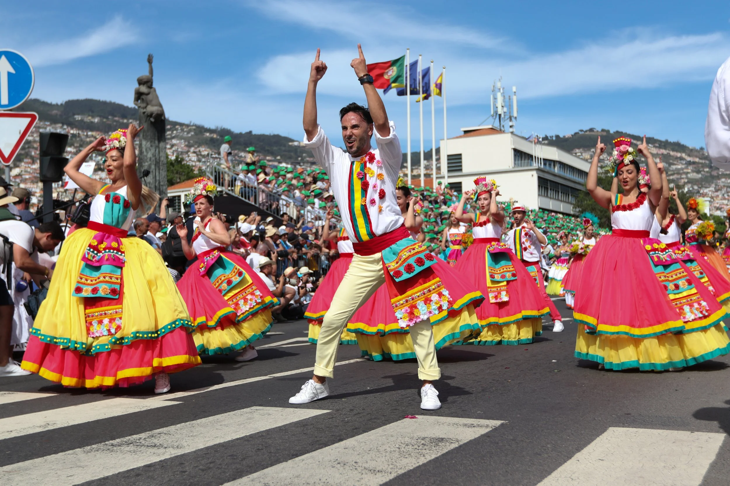 People in colorful traditional costumes dancing during a parade on a city street with spectators, flags, and a hillside background.