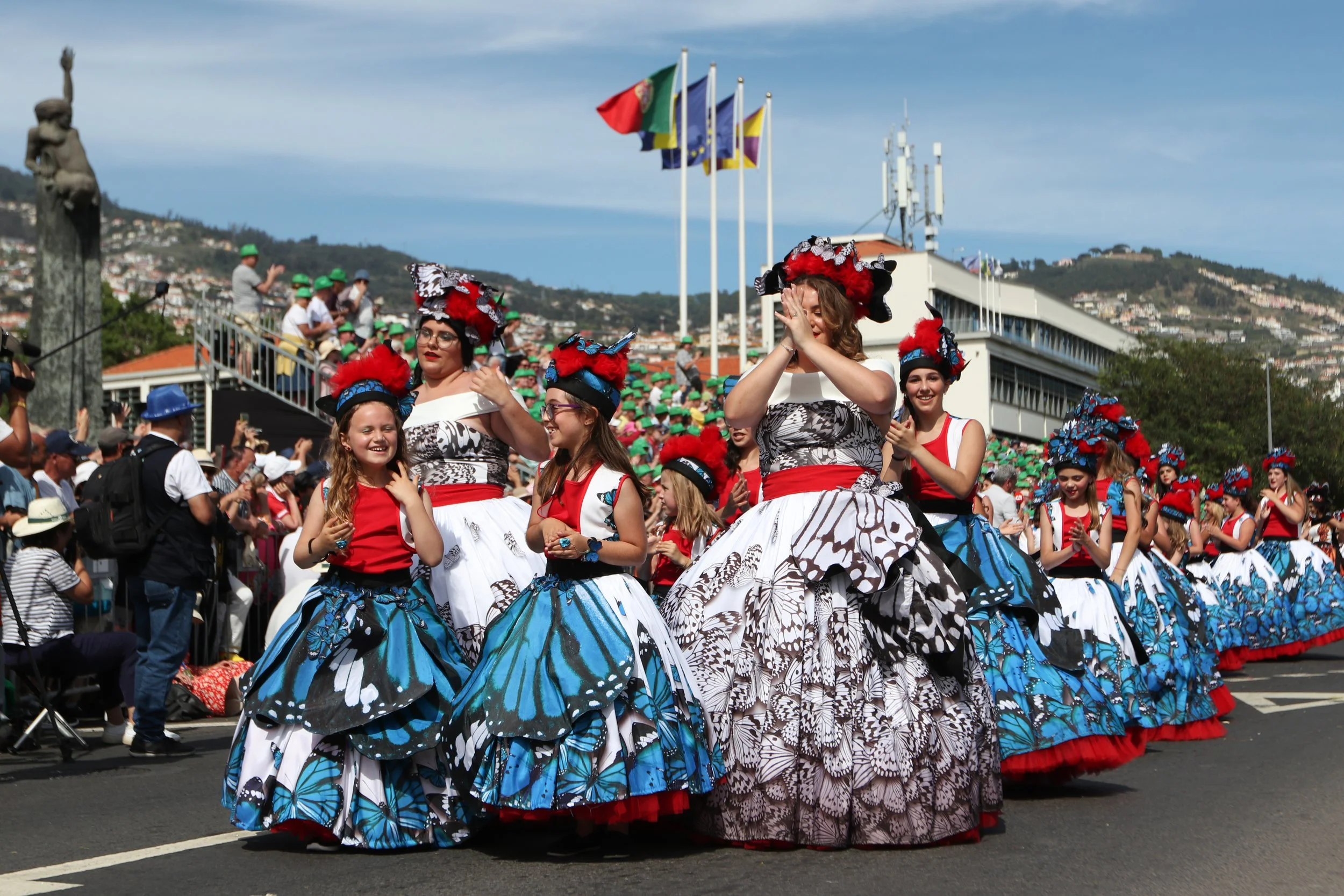 People participating in a parade, dressed in colorful, butterfly-themed costumes with floral patterns and elaborate hats decorated with feathers and flowers, waving and smiling as they march on a city street with flags flying in the background.