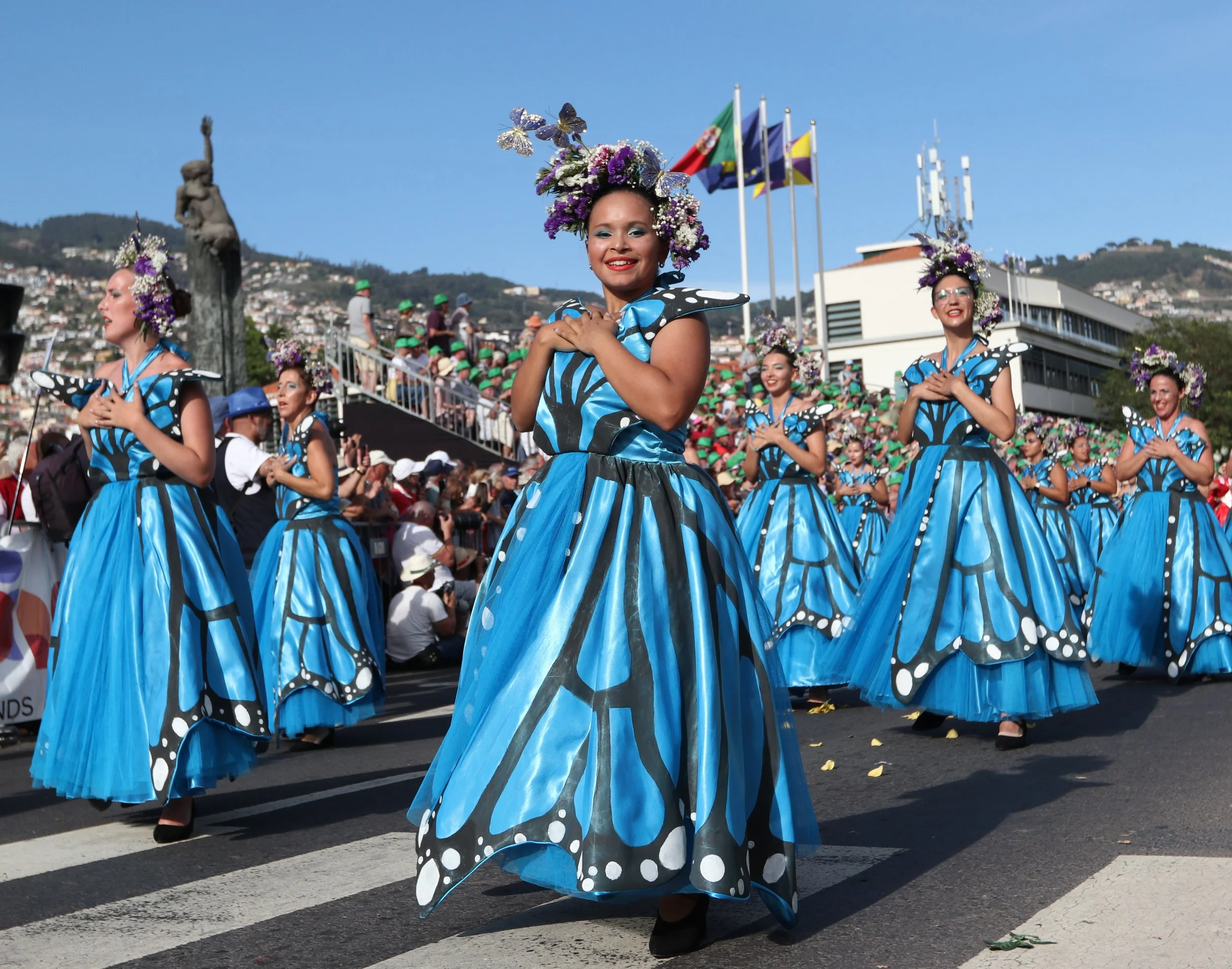 Group of women in blue butterfly costumes with floral headpieces participating in a parade on a city street.