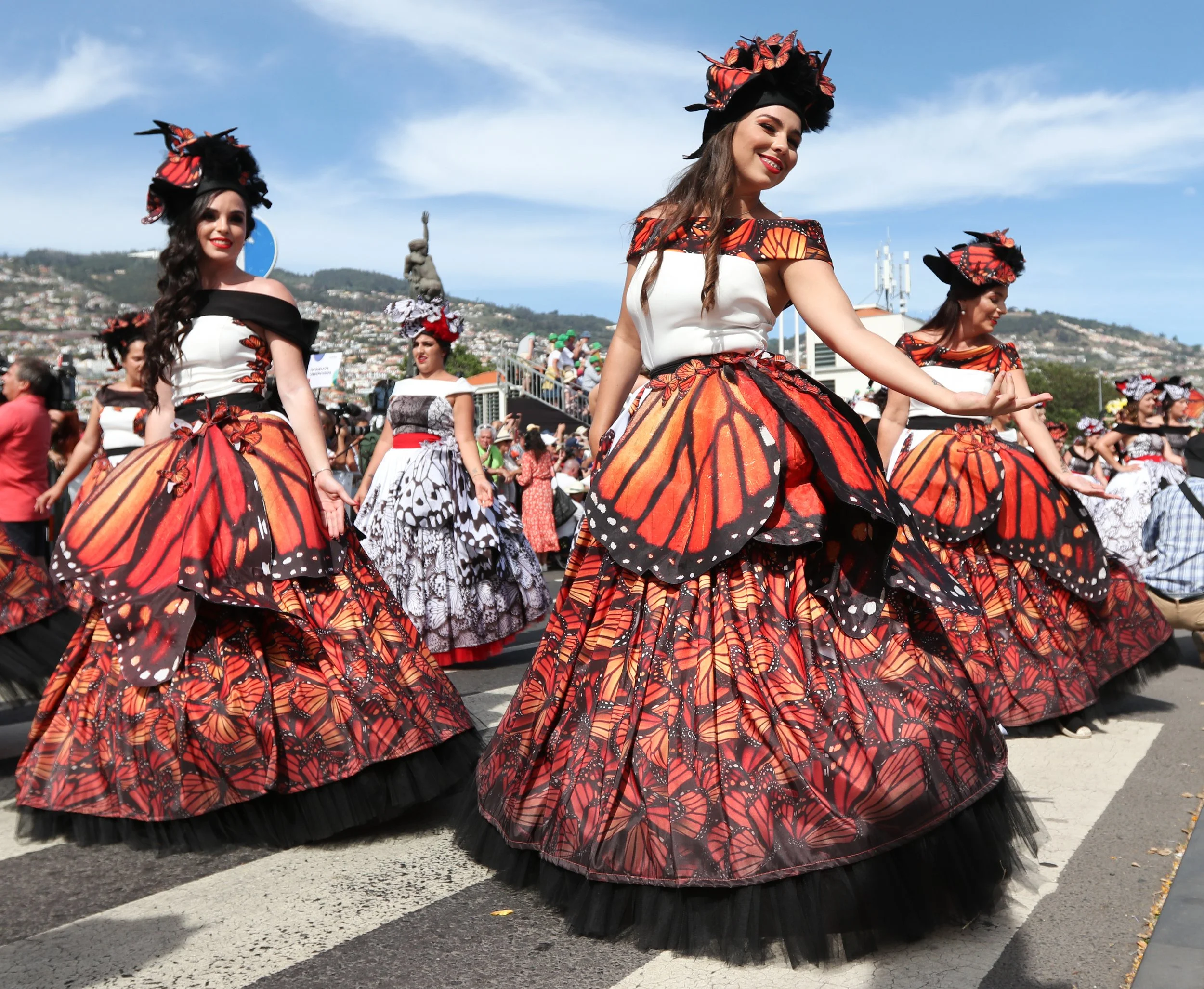 Women in butterfly-themed dresses participating in a parade, with one woman smiling and pointing, and others with similar butterfly skirt designs, on a city street with spectators and hills in the background.