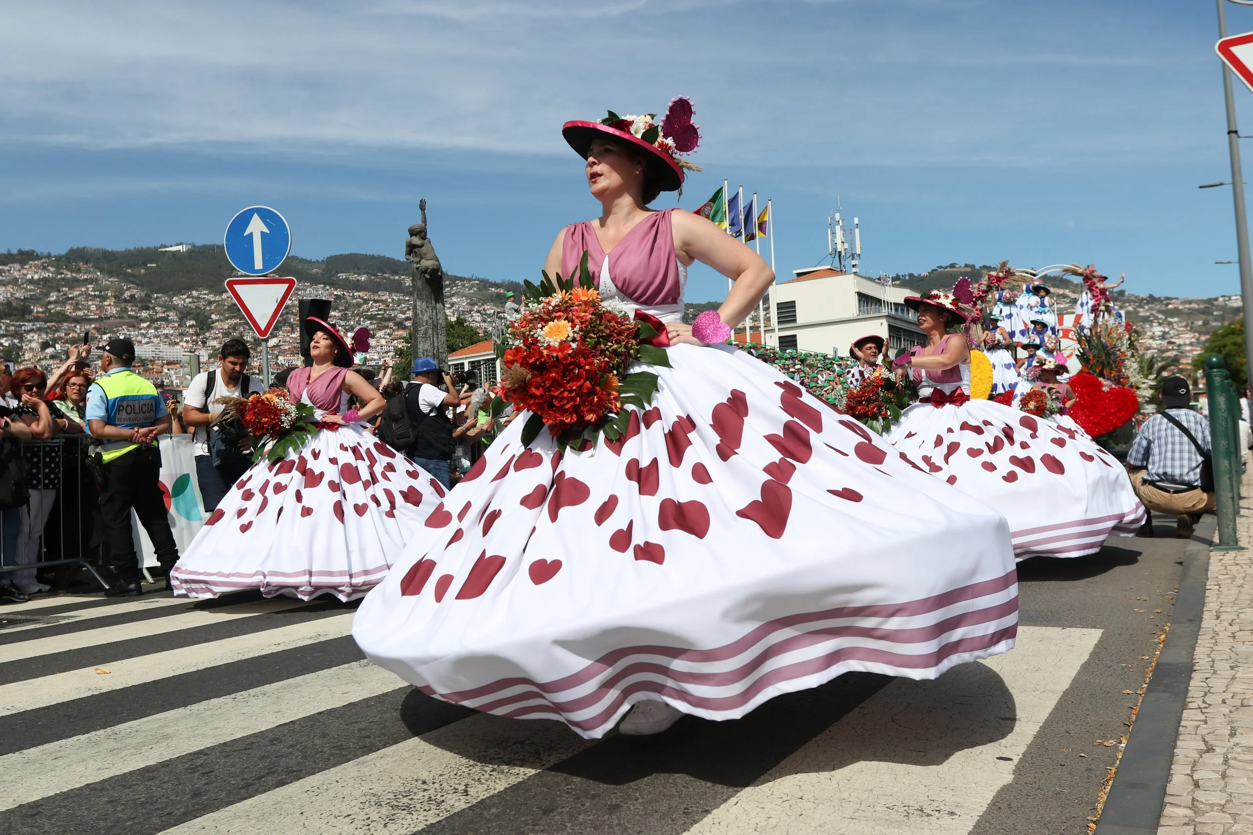 Women in colorful, floral dress costumes participating in a parade on a street, with onlookers and police officers watching. The women are wearing large, decorated hats and holding bouquets of flowers.