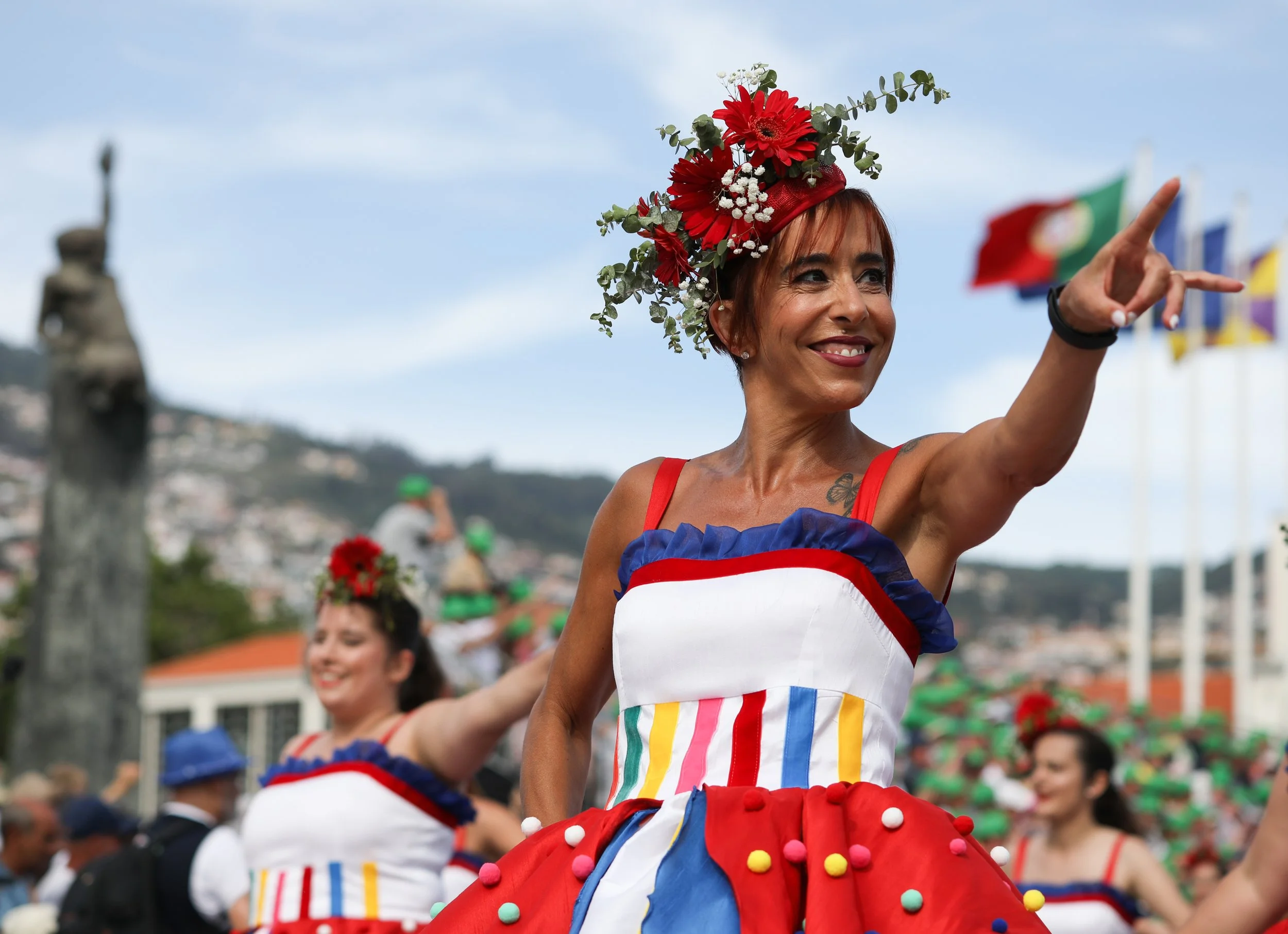 Woman in colorful dress and floral headpiece smiling and pointing during a parade, with other women in similar attire in the background and flags fluttering.