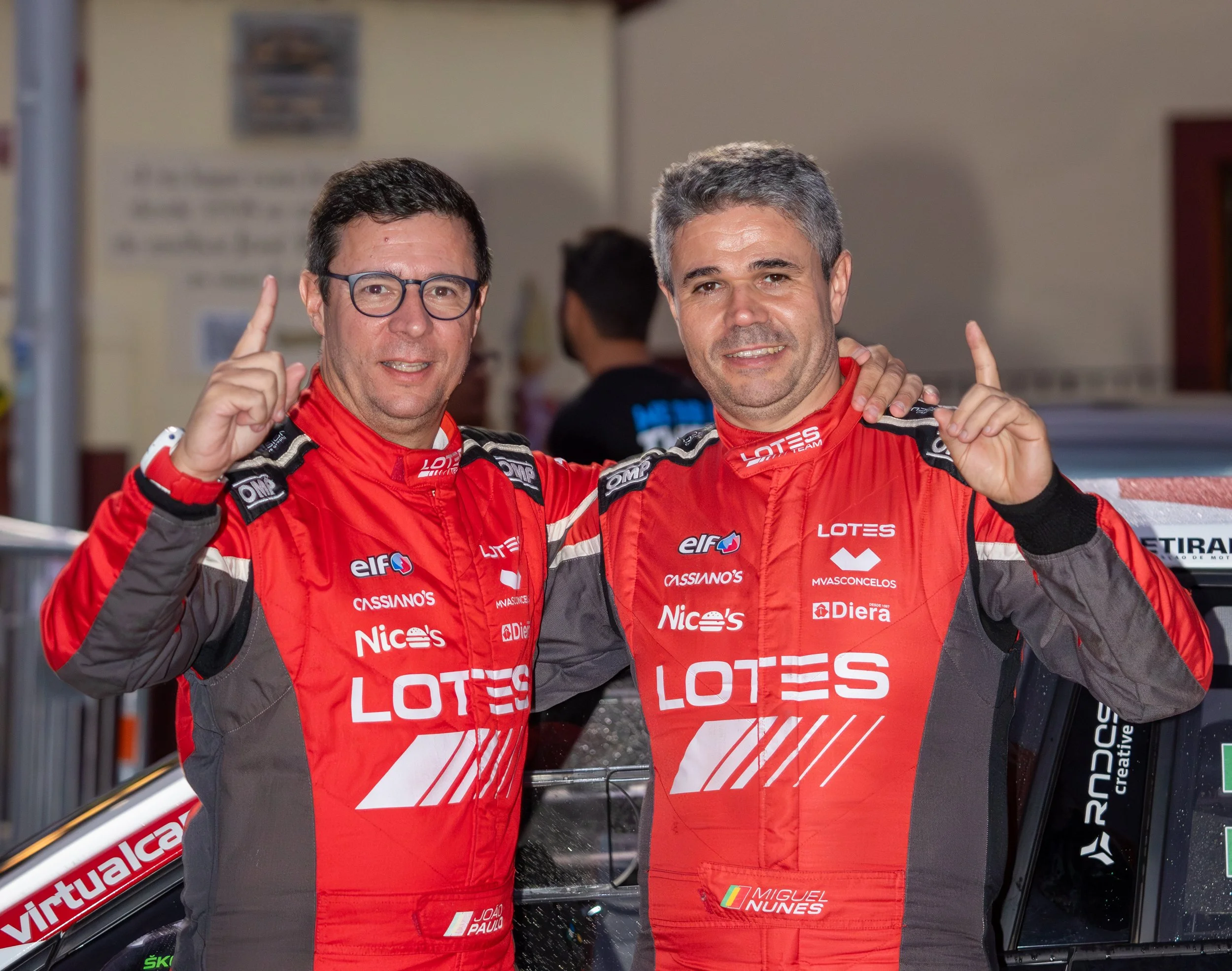 Two race car drivers in red and black racing suits, standing beside a car, smiling and pointing upwards with one finger.