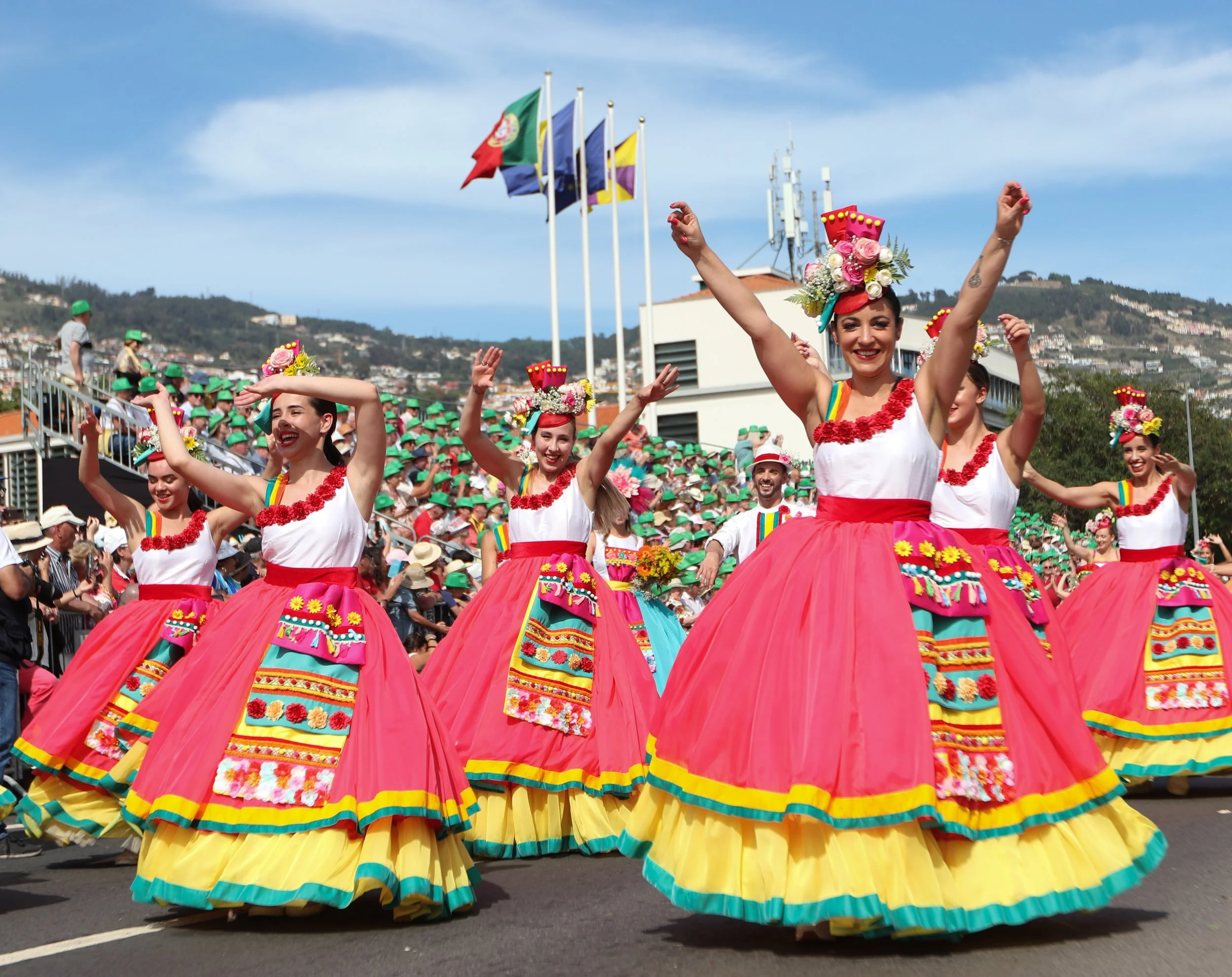 Women dressed in colorful traditional dresses dancing in a parade with flags and audience in the background.