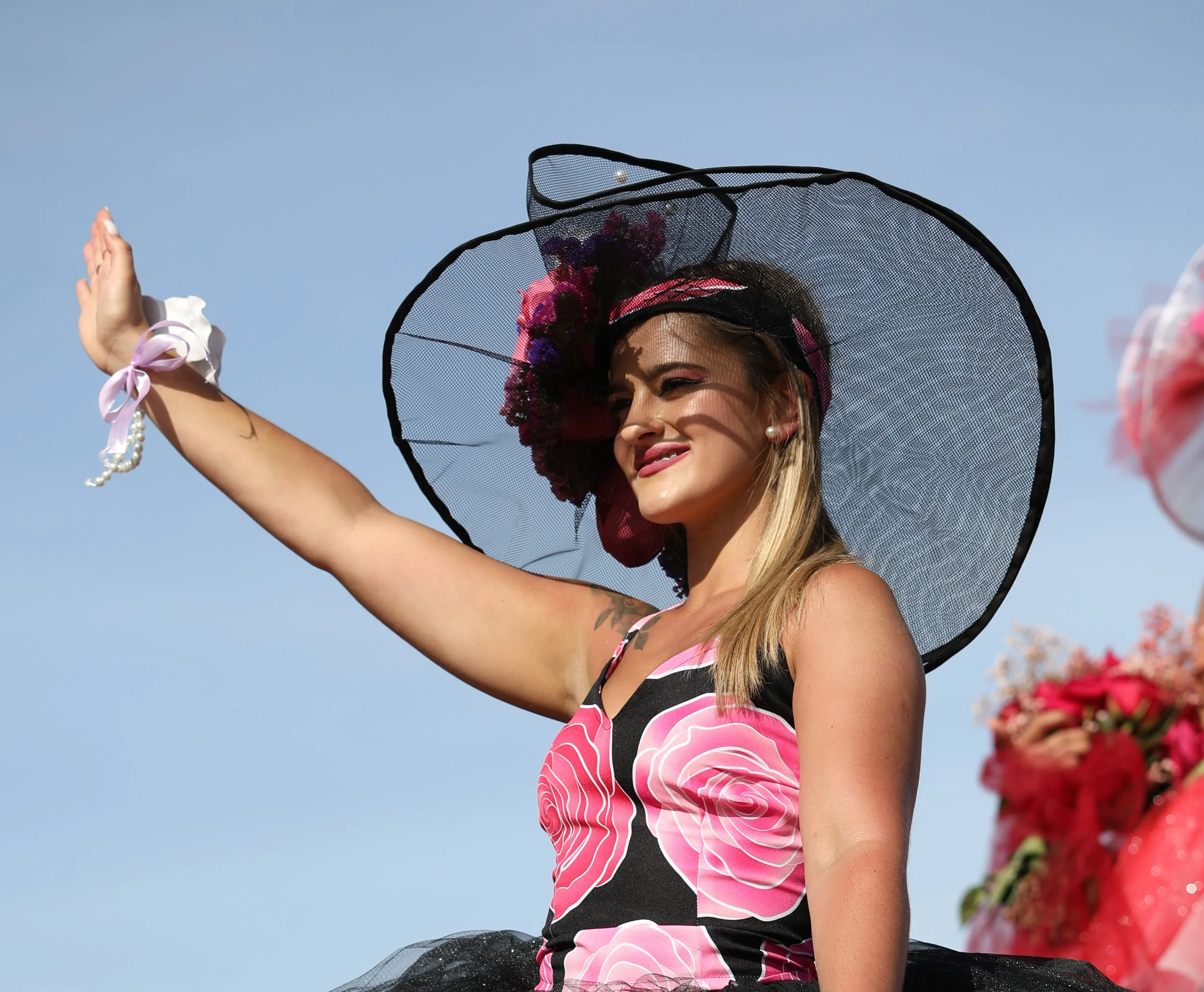 A woman wearing a large black and pink hat, a floral dress, and a bracelet with pearls and ribbons, standing outdoors against a blue sky, smiling and waving.