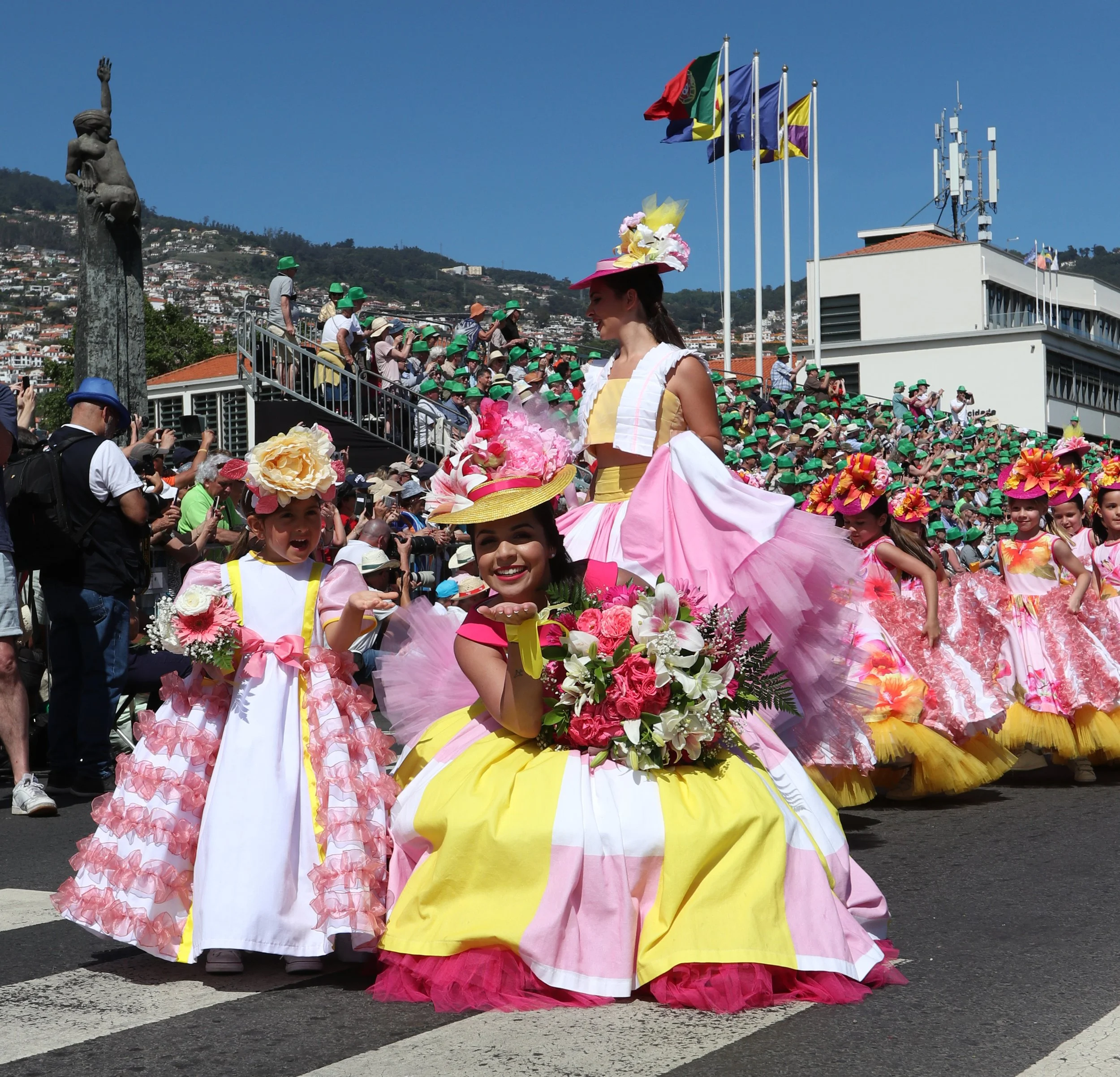 Women and children in colorful floral dresses and hats participating in a parade, with a crowd and flags in the background.