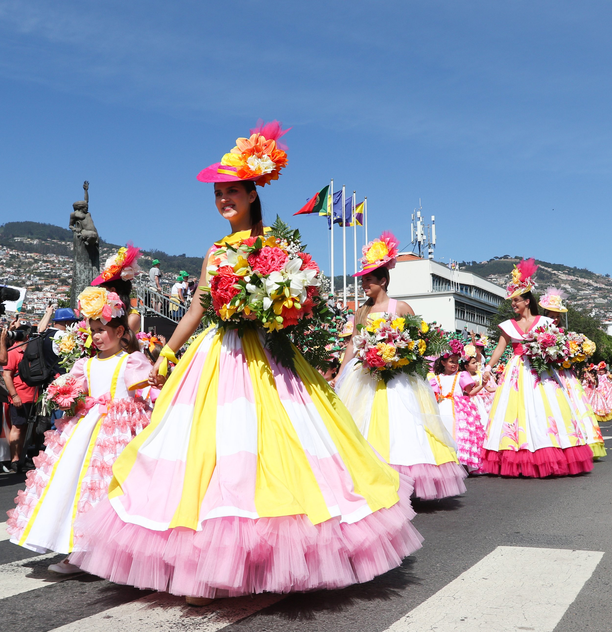 Women and girls in colorful floral dresses and flower hats participating in a parade on a sunny day.