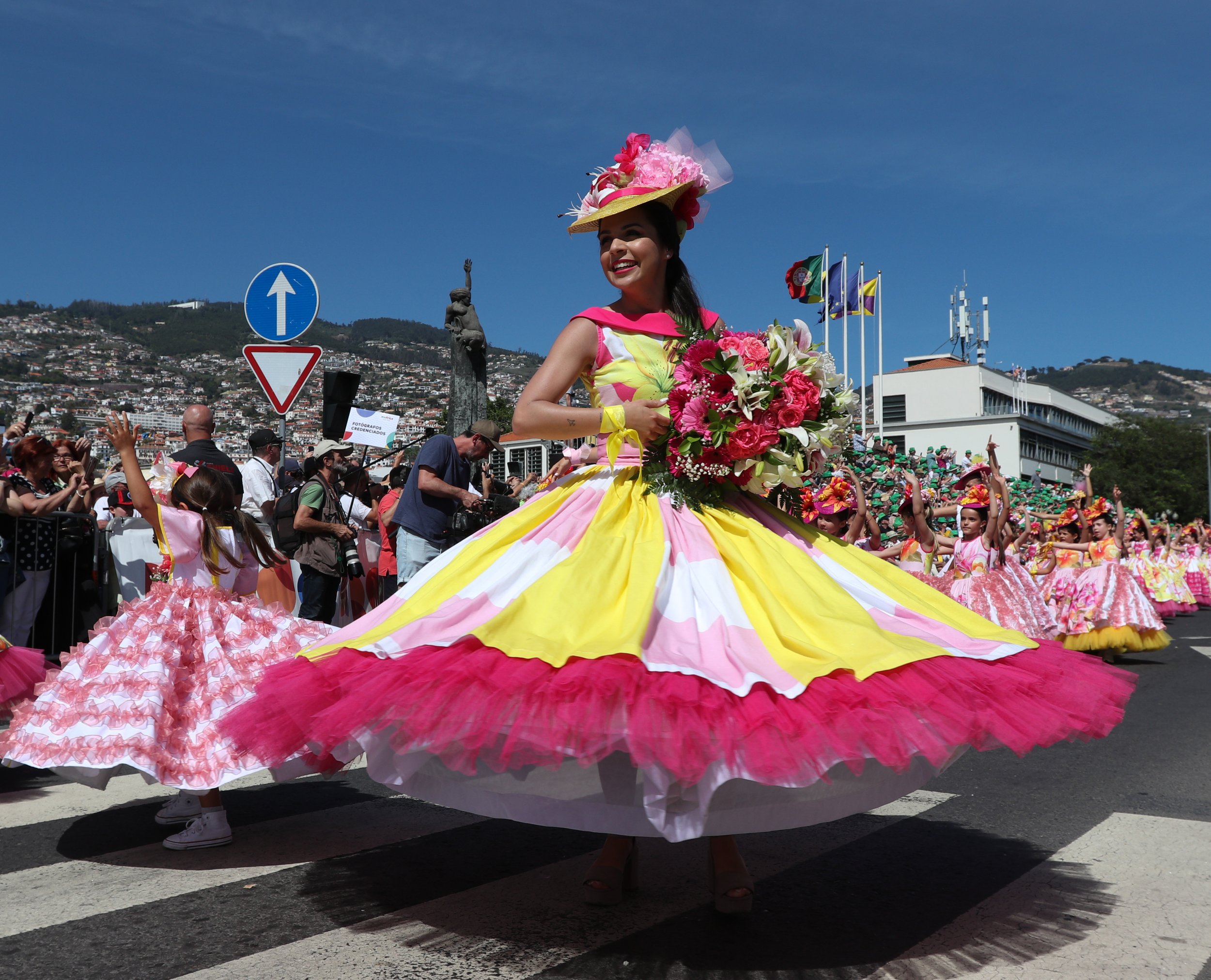 A woman dressed in a colorful, wide, floral-patterned dress with pink and yellow hues, holding a bouquet of pink and white flowers, participating in a parade under a clear blue sky, with children and spectators around.