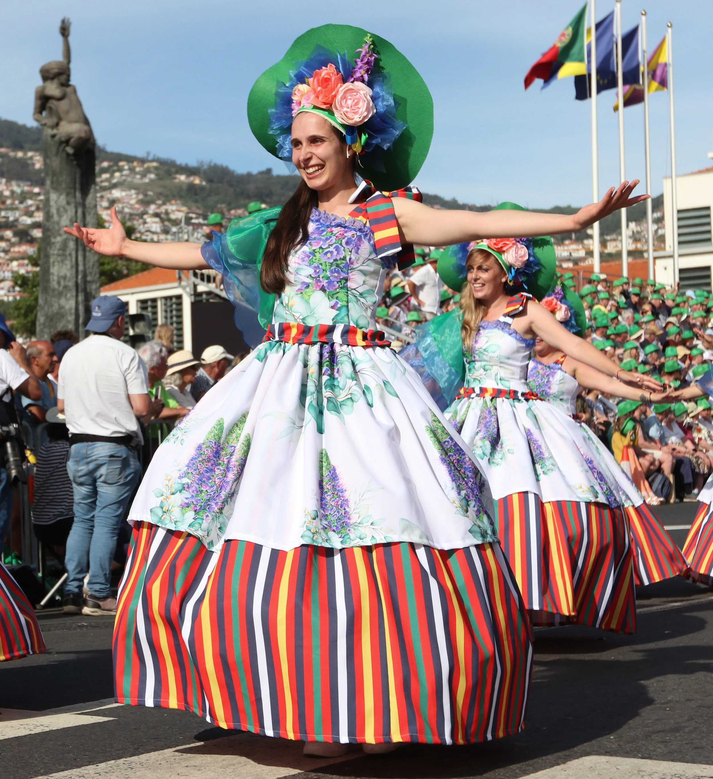 Women wearing colorful floral dresses and large hats performing a traditional dance during a parade with bystanders and flag banners in the background.