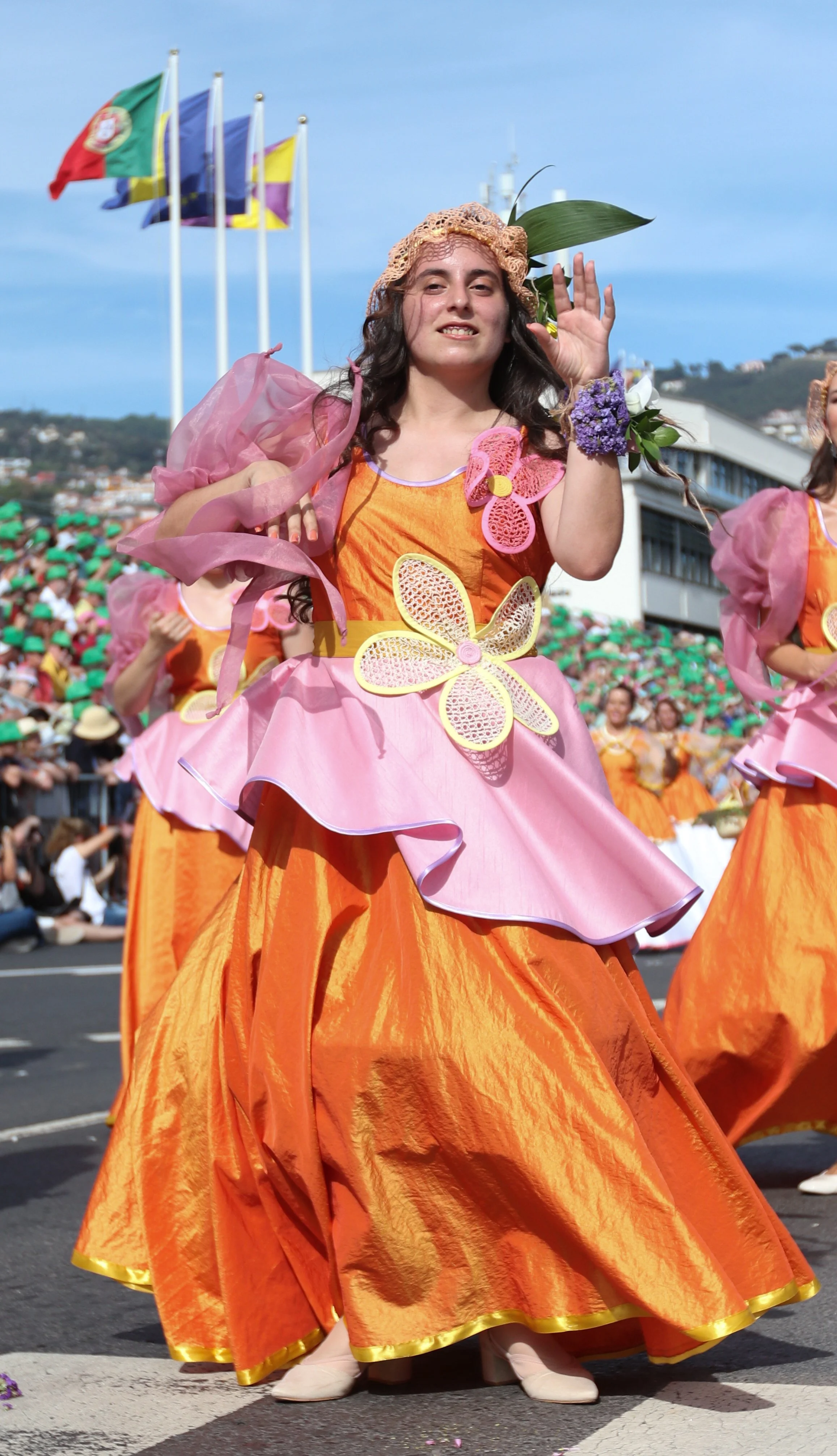 A woman dressed in an orange and pink floral-themed costume participates in a parade. She is waving and smiling, with a crowd and multiple flags in the background.