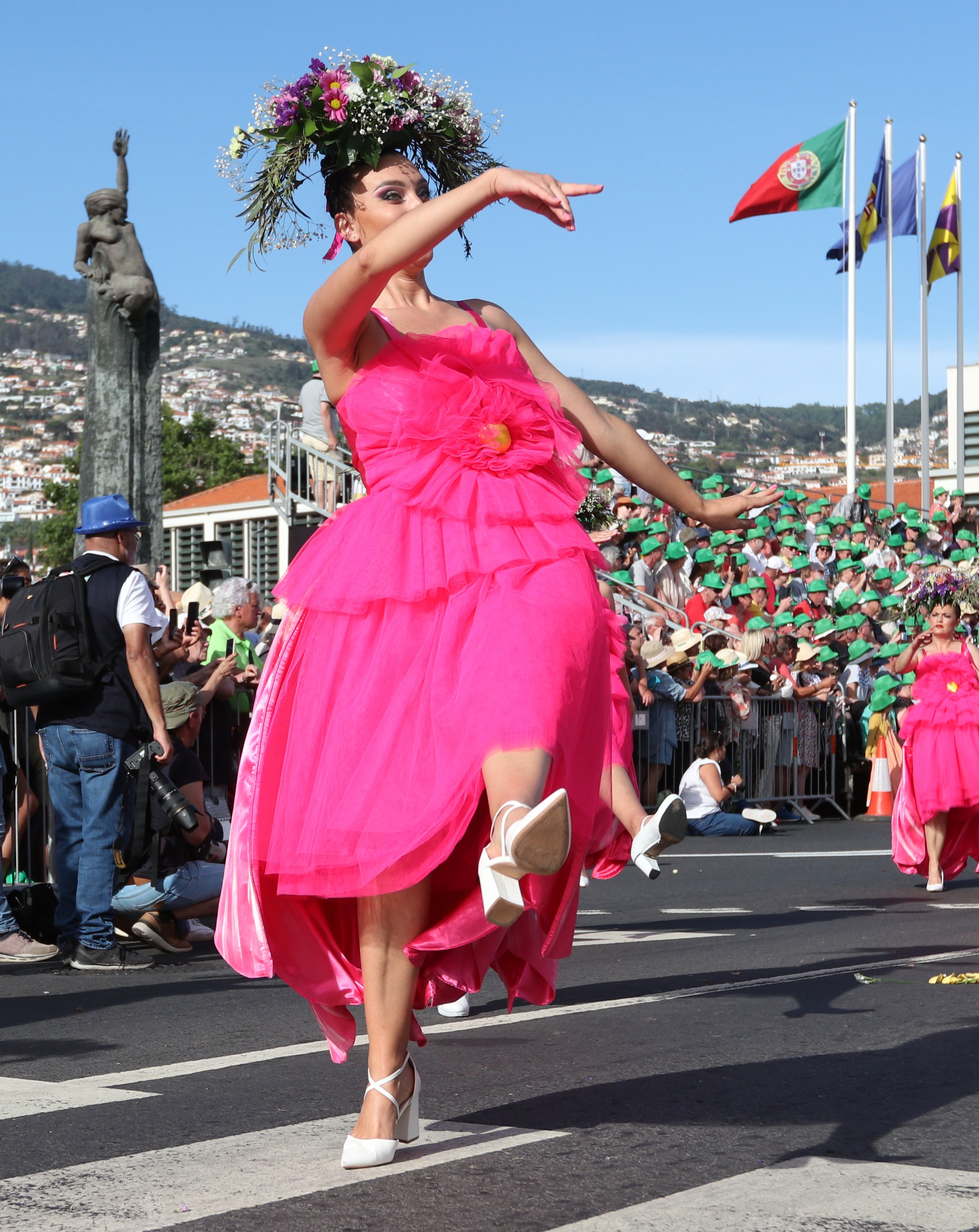A woman in a bright pink dress and floral headpiece dancing in a parade with spectators and flags in the background.
