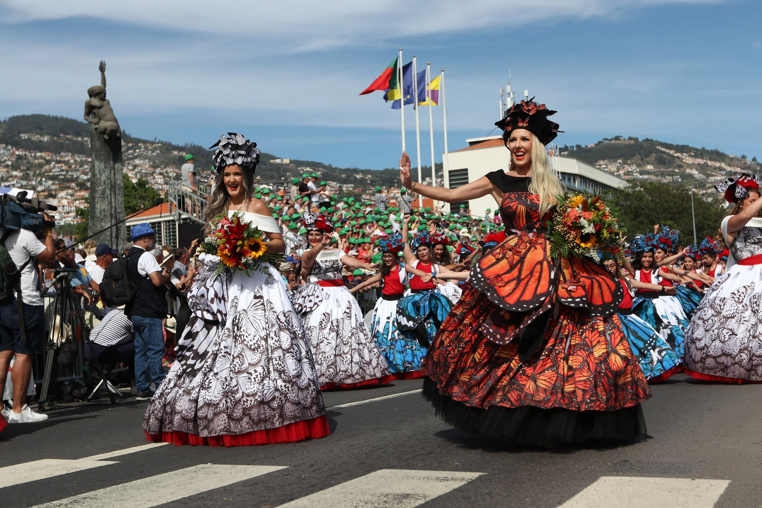 Colorful parade featuring women in vibrant flamenco-style skirts with butterfly and floral designs, participating in a festival on a city street, with flags and a crowd of spectators in the background, hills, and cityscape visible.