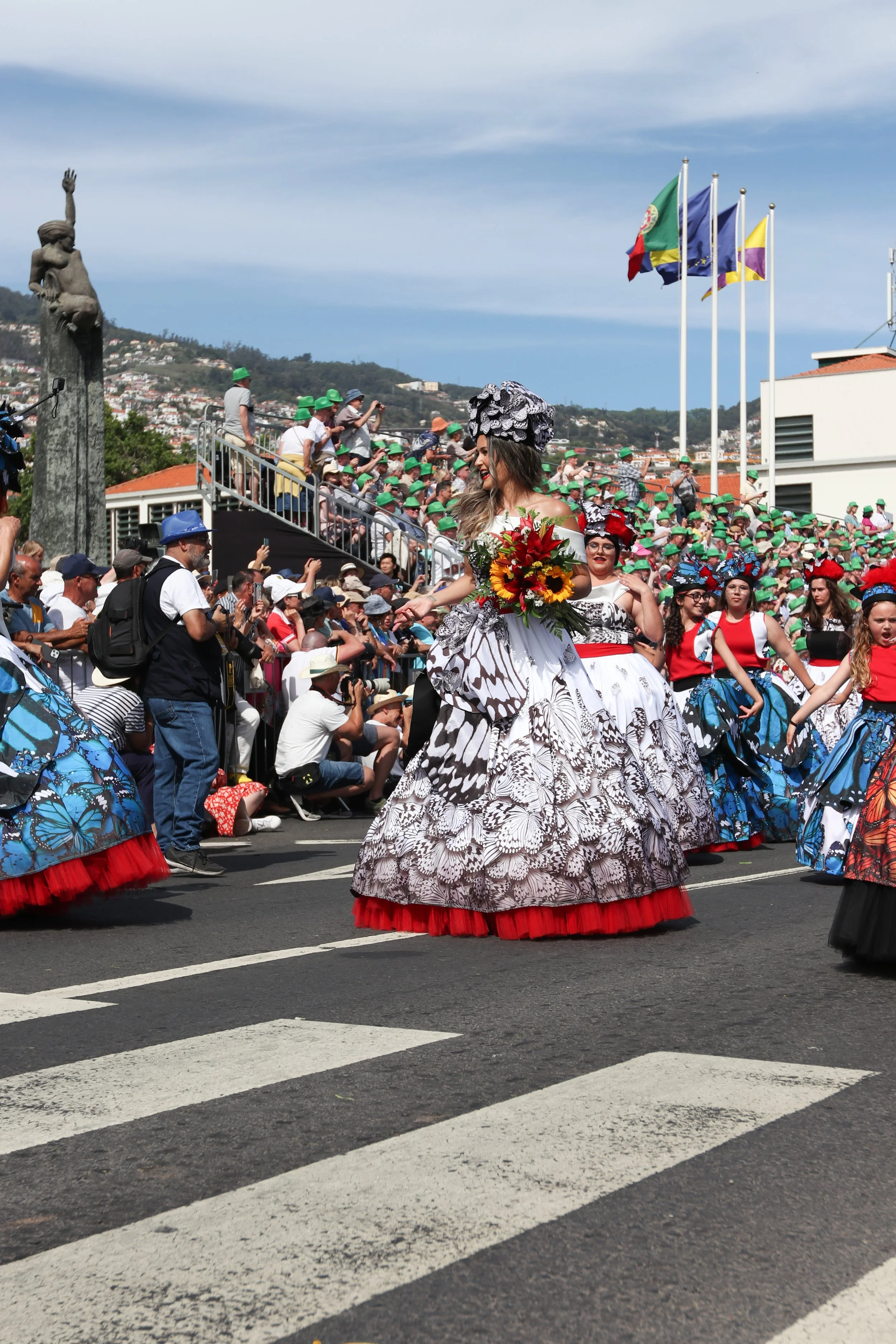 Women dressed in colorful traditional costumes with butterfly patterns walking in a parade, holding a bouquet of flowers, with spectators watching from the sidewalk and a hillside in the background.