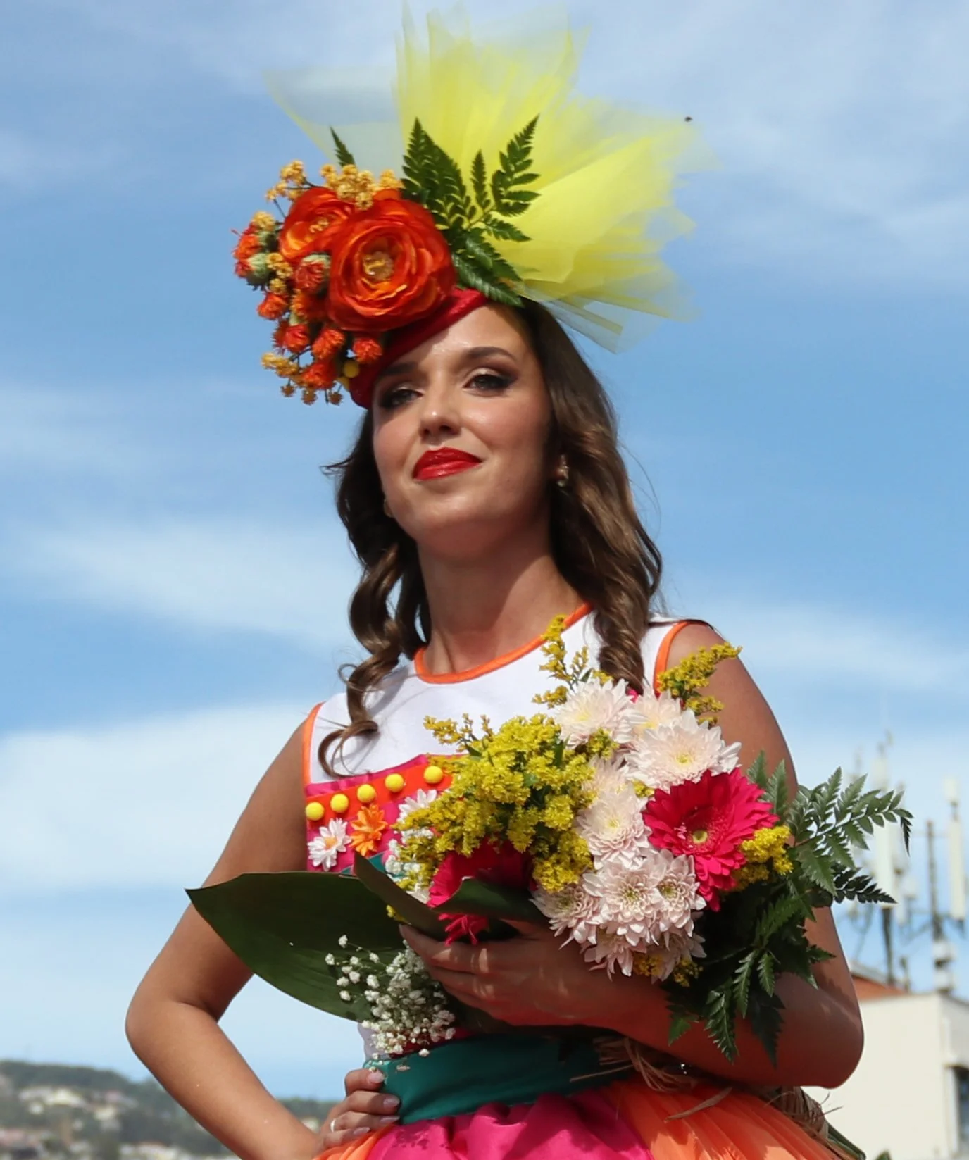 Woman wearing a floral headpiece and colorful dress holding a bouquet of flowers outdoors against blue sky.