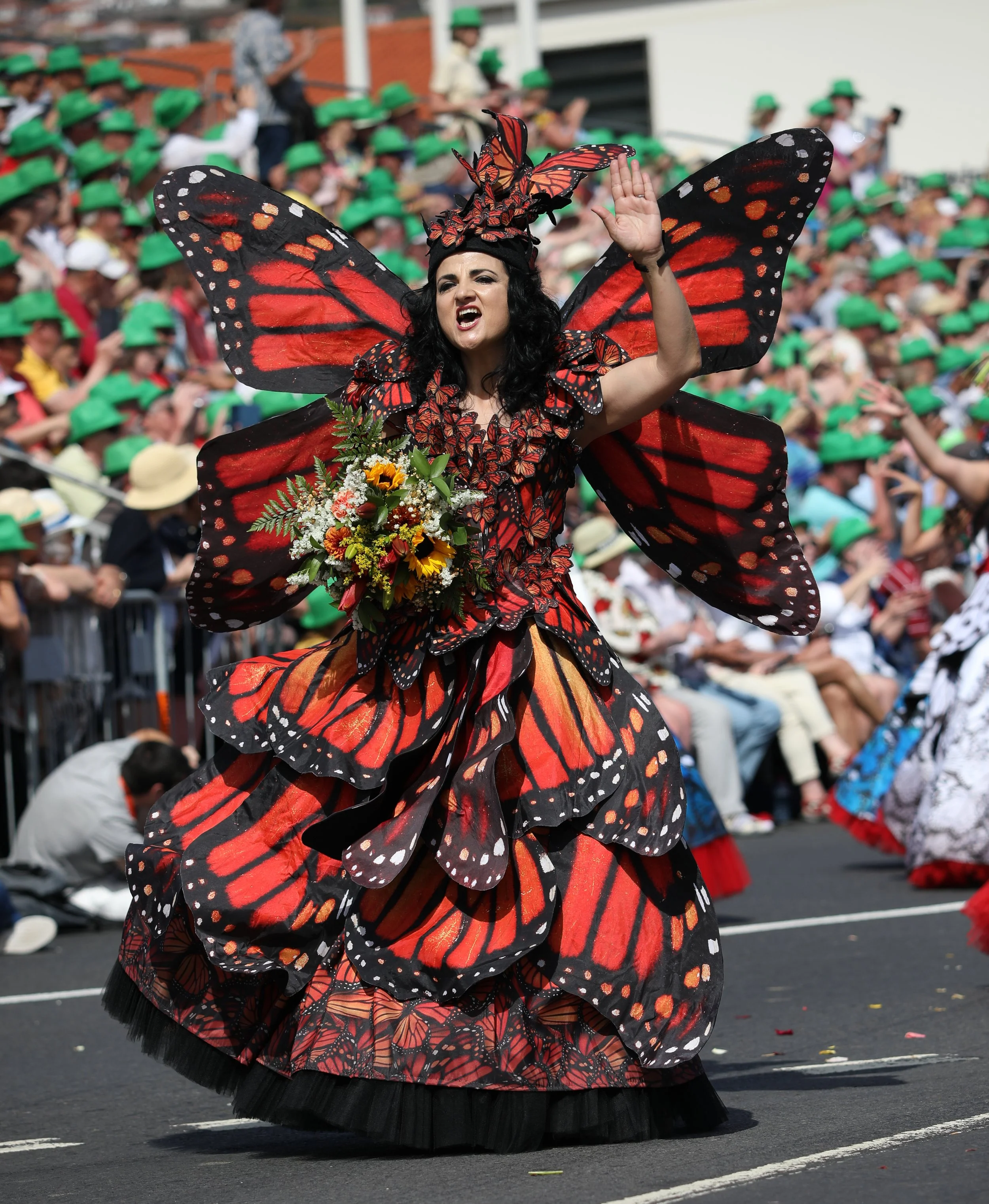 A woman dressed as a butterfly with large orange and black wings and a matching dress, holding a bouquet of flowers, participating in a parade with a crowd of people wearing green hats in the background.
