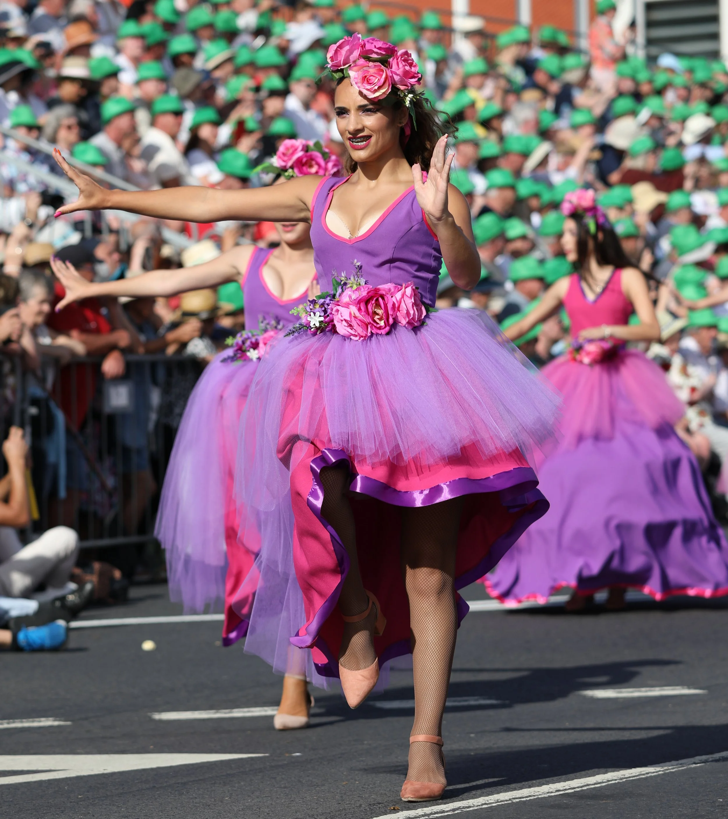 Women in colorful purple and pink dresses with floral accessories participate in a parade on a city street, with spectators watching from the sidelines.
