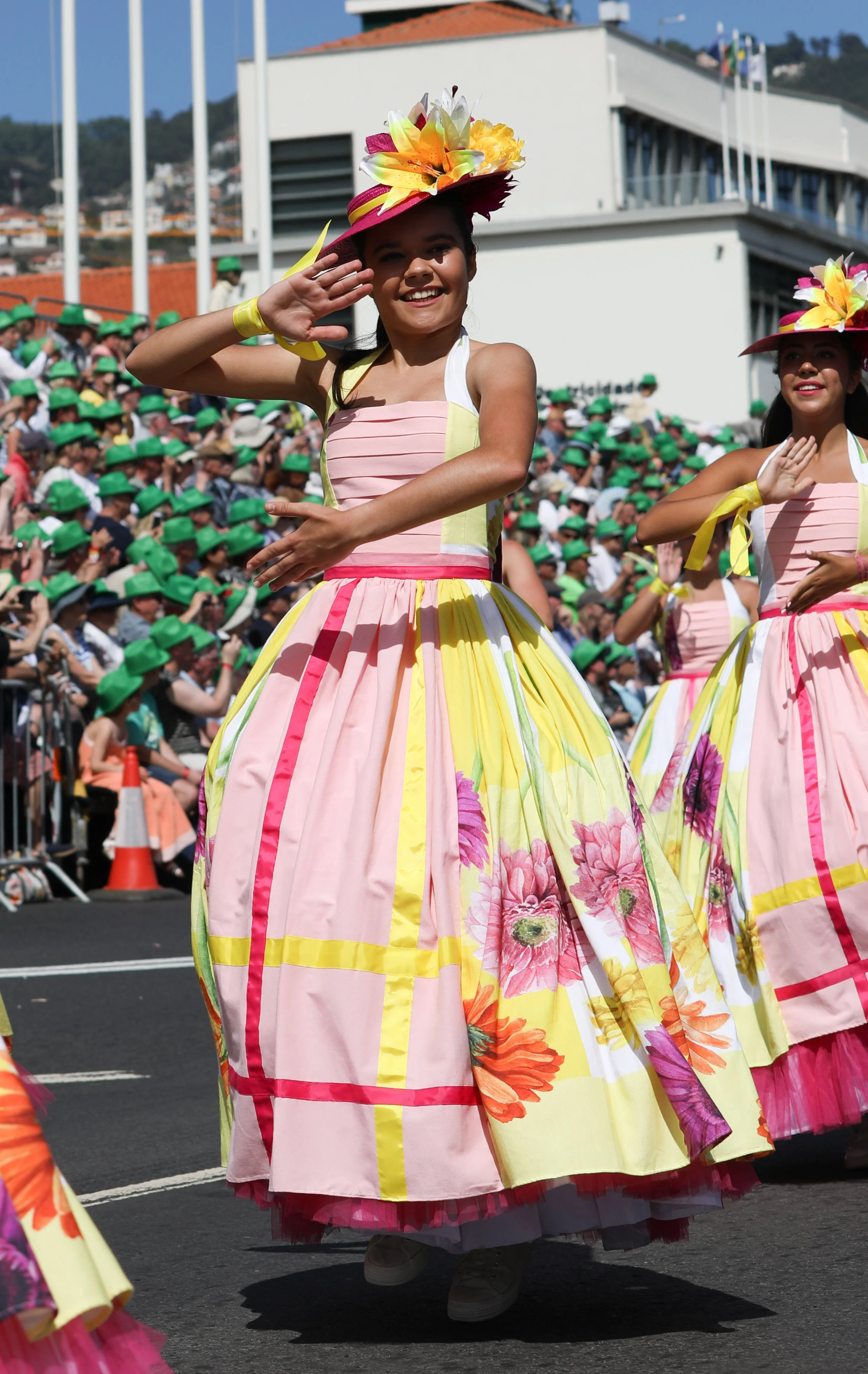 Women in colorful floral dresses and large floral hats dancing in a parade, with a crowd of spectators wearing green hats in the background.