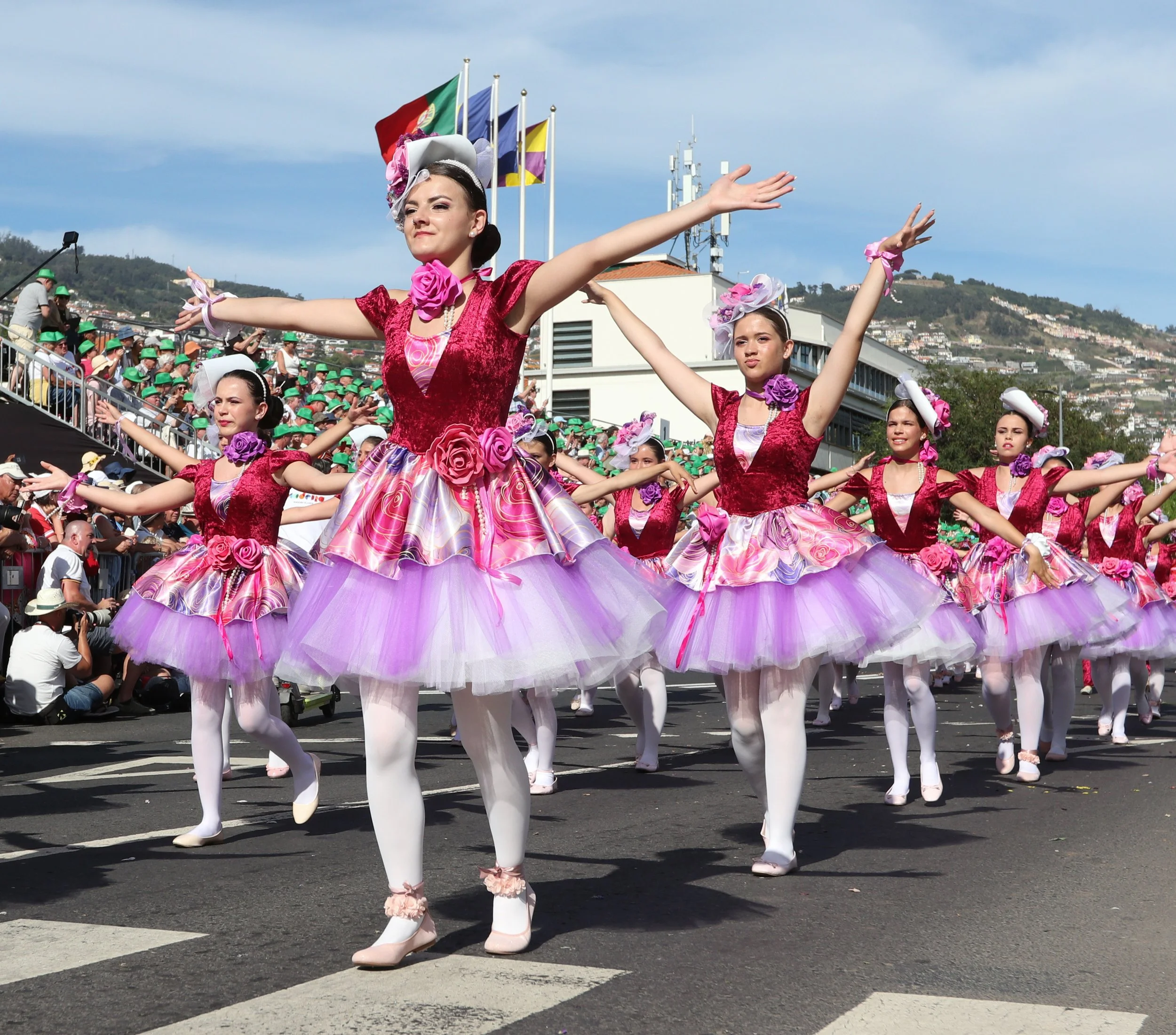 Group of women in pink and purple tutus dancing in a parade on a city street, spectators watching from the sidelines.