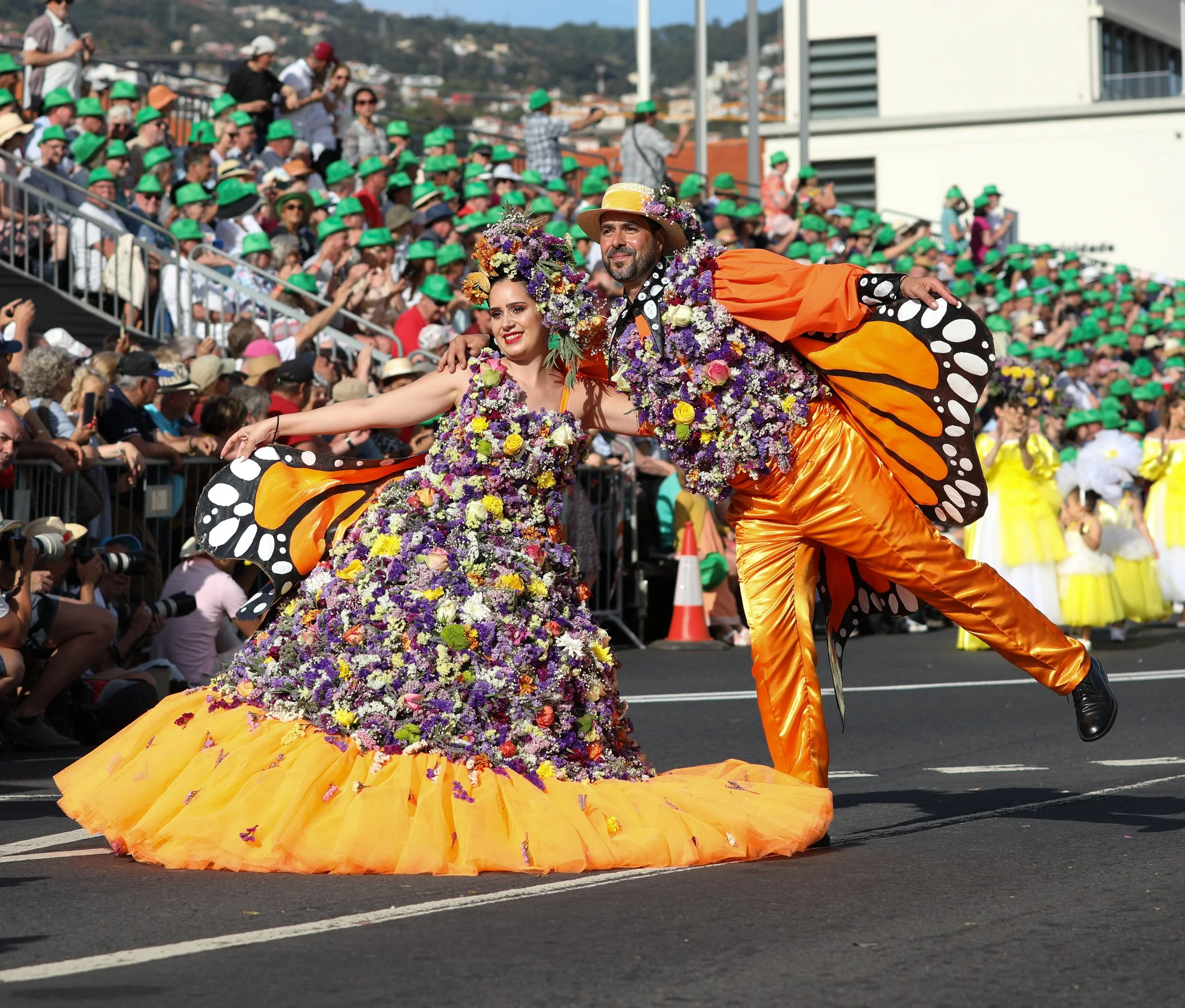 Two performers dressed in colorful costumes with butterfly wings and flower embellishments dancing in a parade. A large crowd with many wearing green hats watches from the crowd.