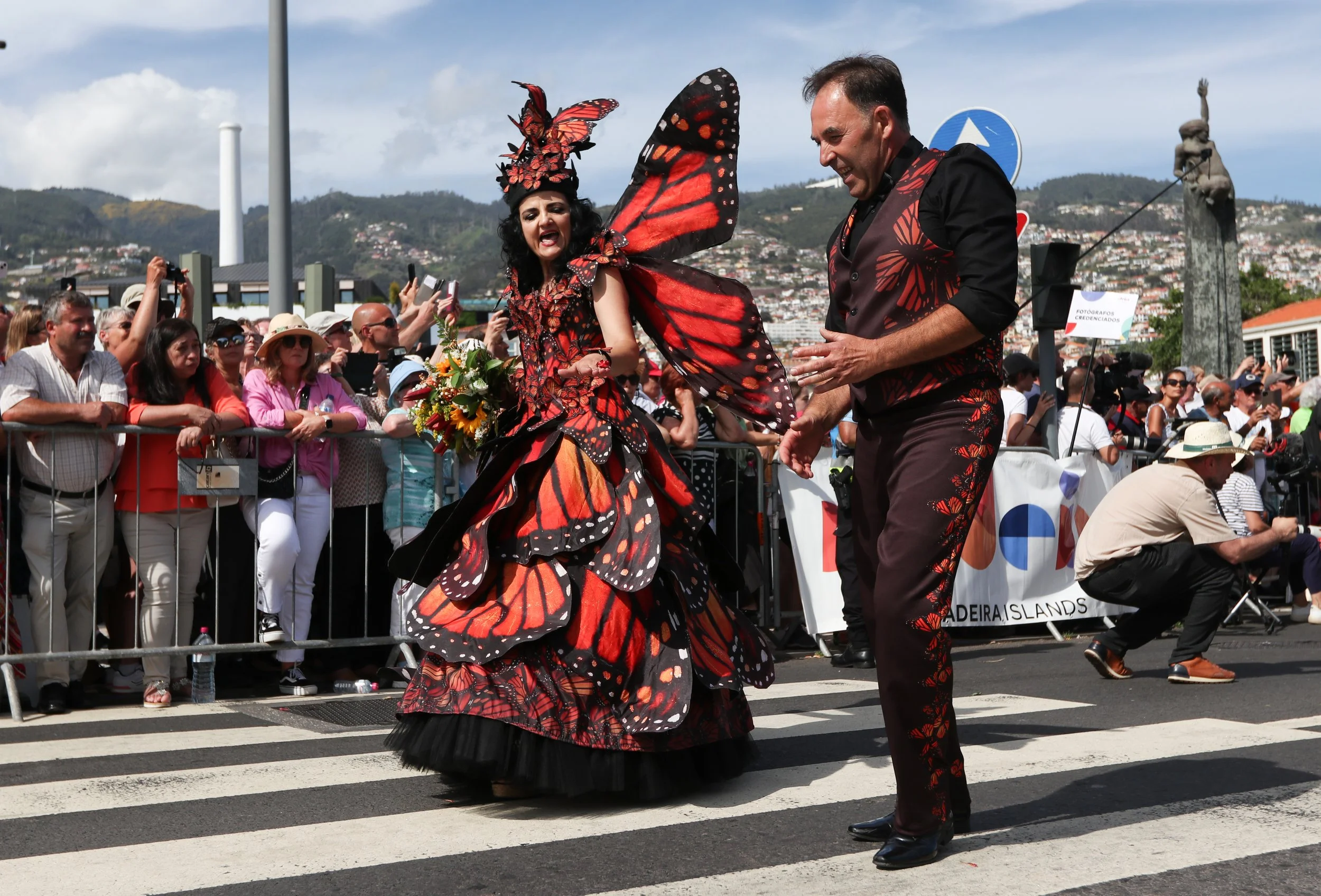A woman dressed as a butterfly with butterfly wings and a colorful dress is holding a bouquet of flowers, dancing on a street during a parade, with a smiling man in coordinated outfit and a crowd watching behind a barricade.