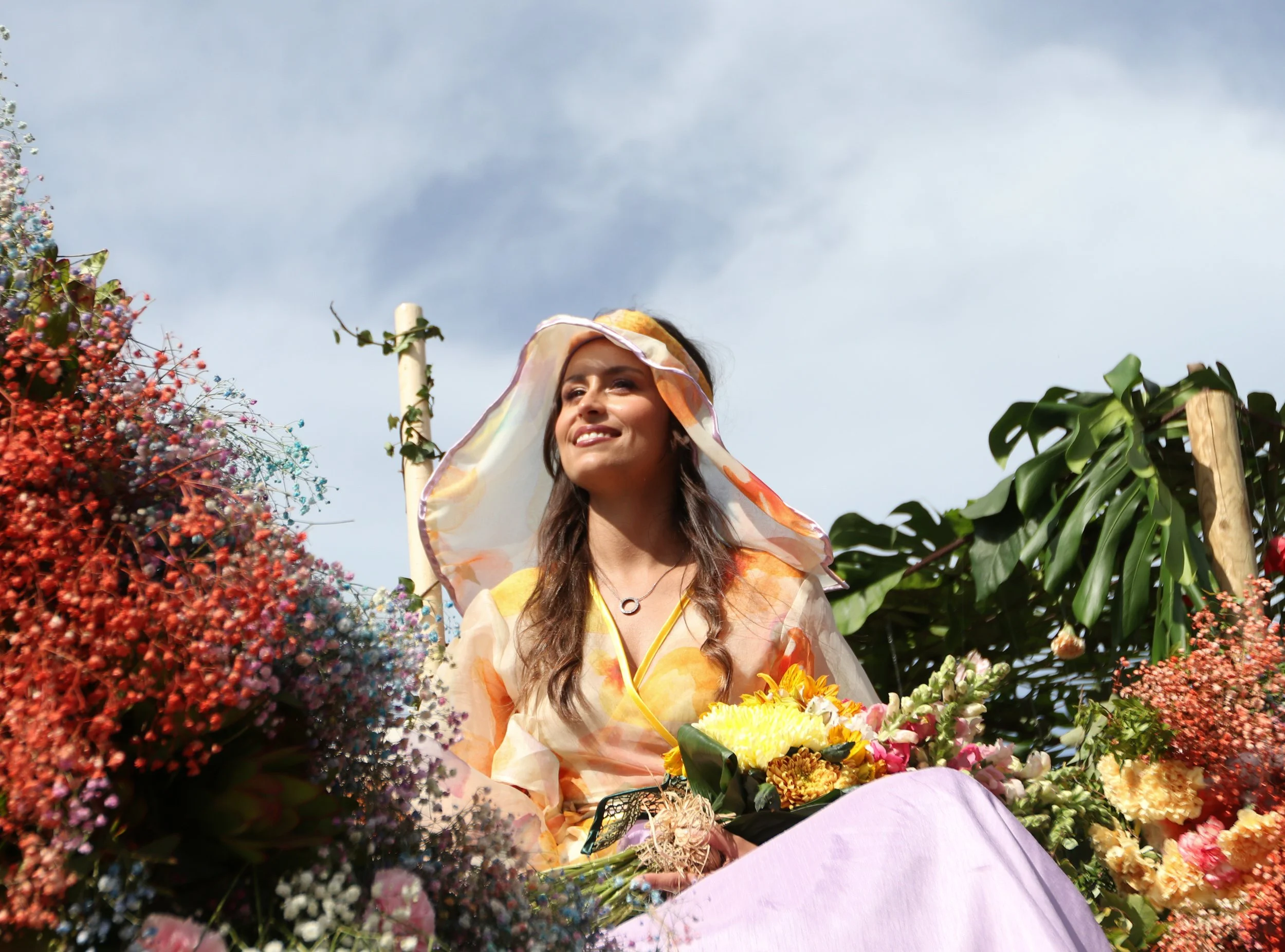 Woman in floral dress with a wide-brimmed hat sitting among colorful flowers outdoors on a sunny day.
