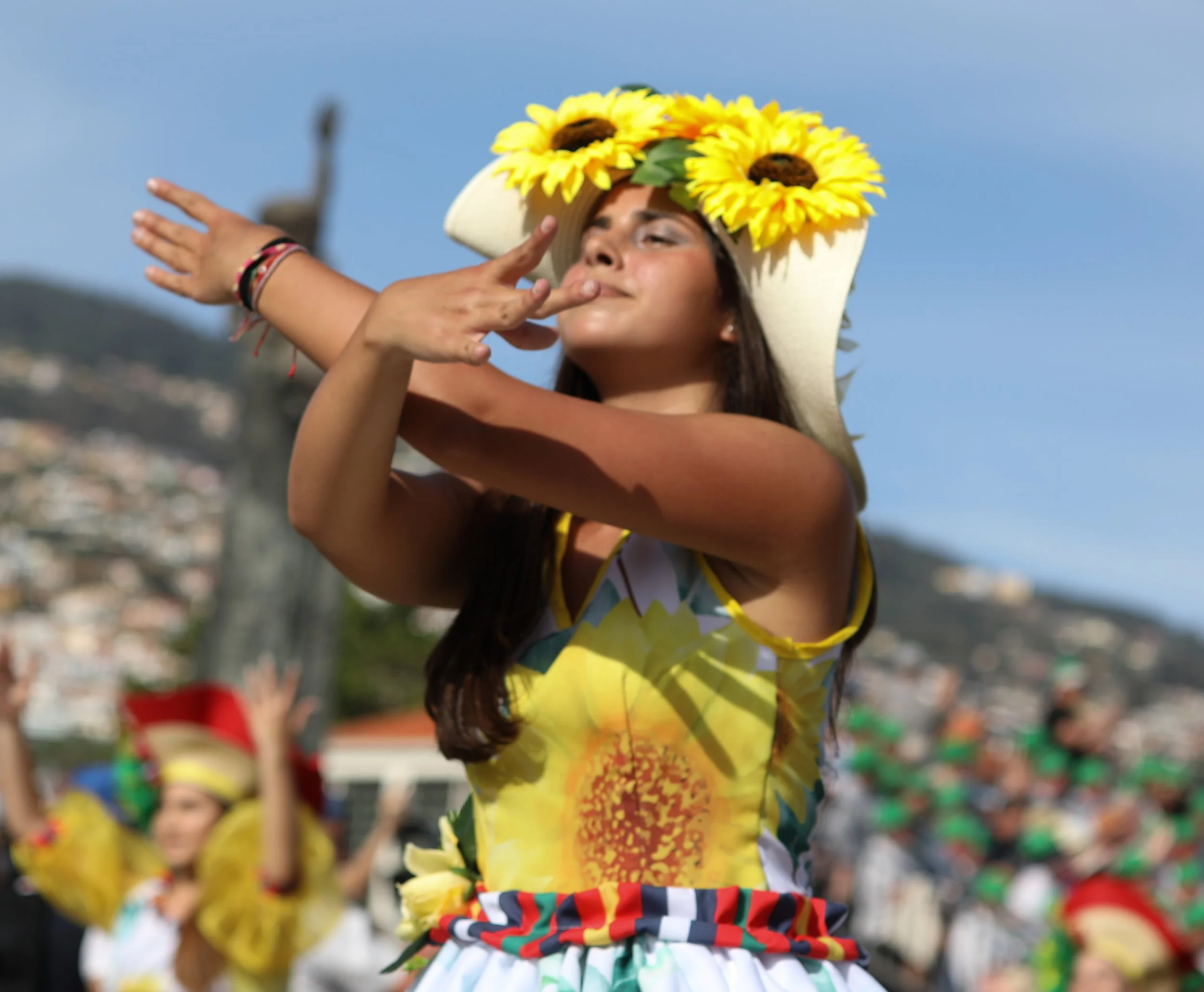 Young woman dancing outdoors during a cultural festival, wearing a sunflower-themed headpiece and a colorful floral dress.