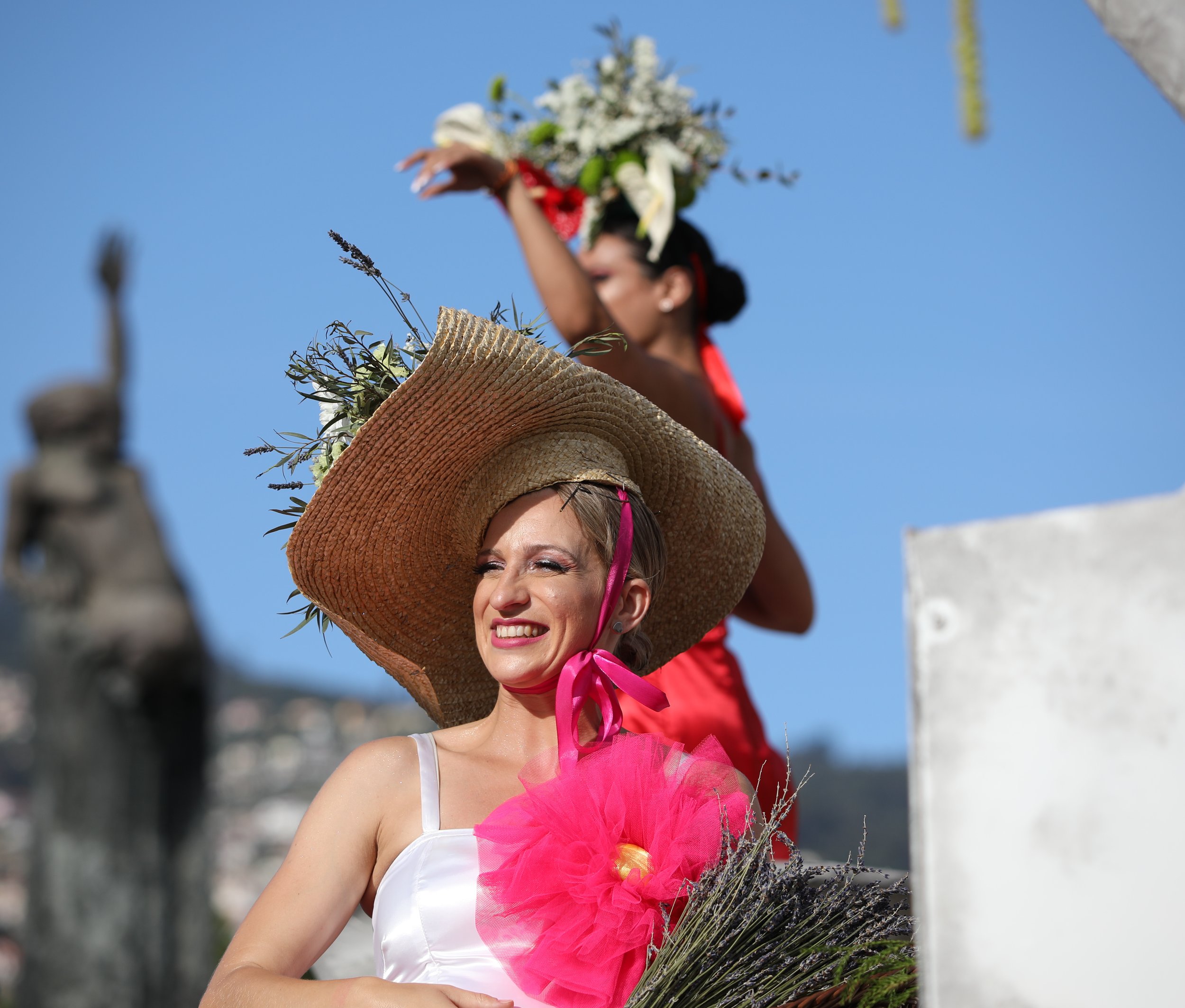 Smiling woman wearing a large wide-brimmed straw hat with flowers and a pink ribbon, holding a bouquet, at an outdoor sunny event, with a woman in the background wearing a floral crown.