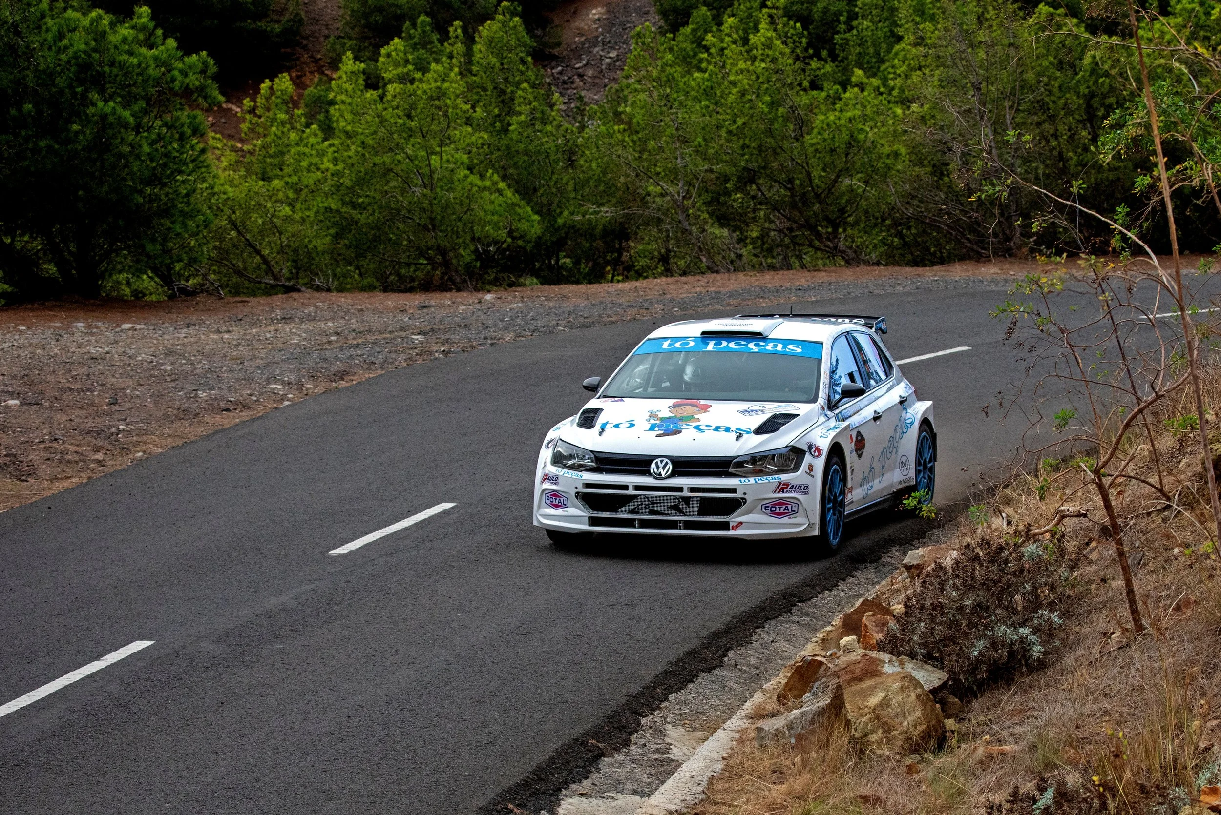 A white rally car with blue accents and various logos, including 'to peças', driving on a winding mountain road surrounded by trees and rocky terrain.