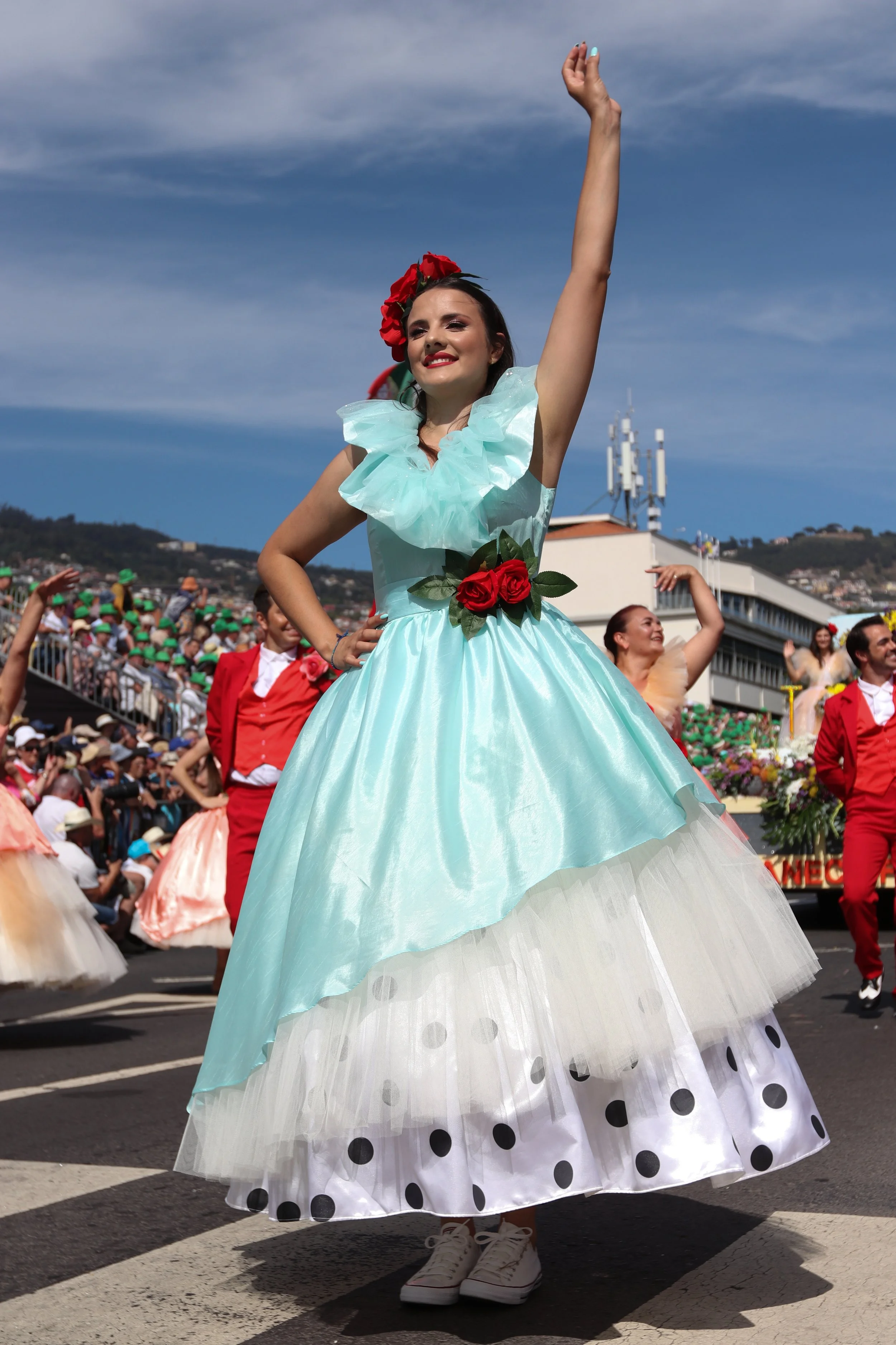 A woman in a colorful dress with a red flower headpiece dancing in a parade with a crowd in the background.