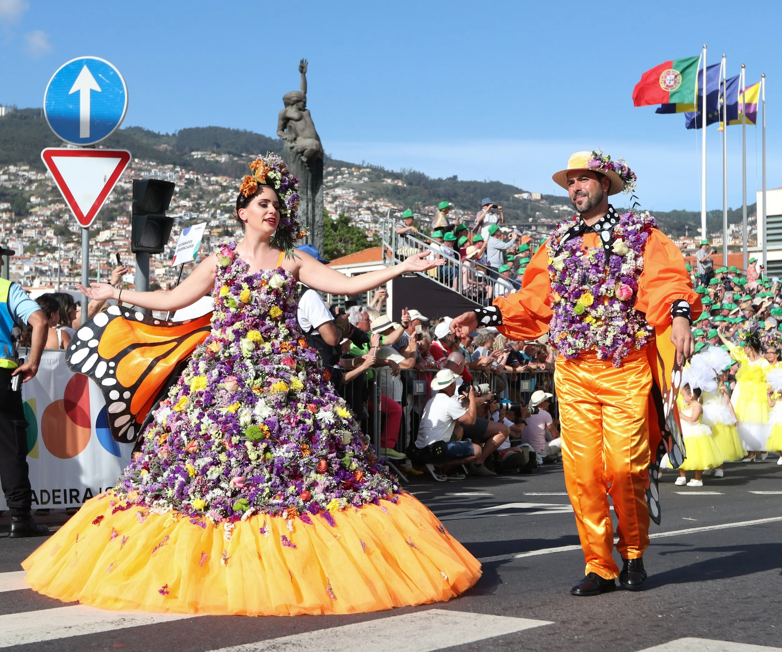 Festival parade with two people dressed in colorful costumes decorated with flowers. The woman wears a large floral dress with butterfly wings and the man wears bright orange pants and floral accessories. Flags are visible in the background, and a cr