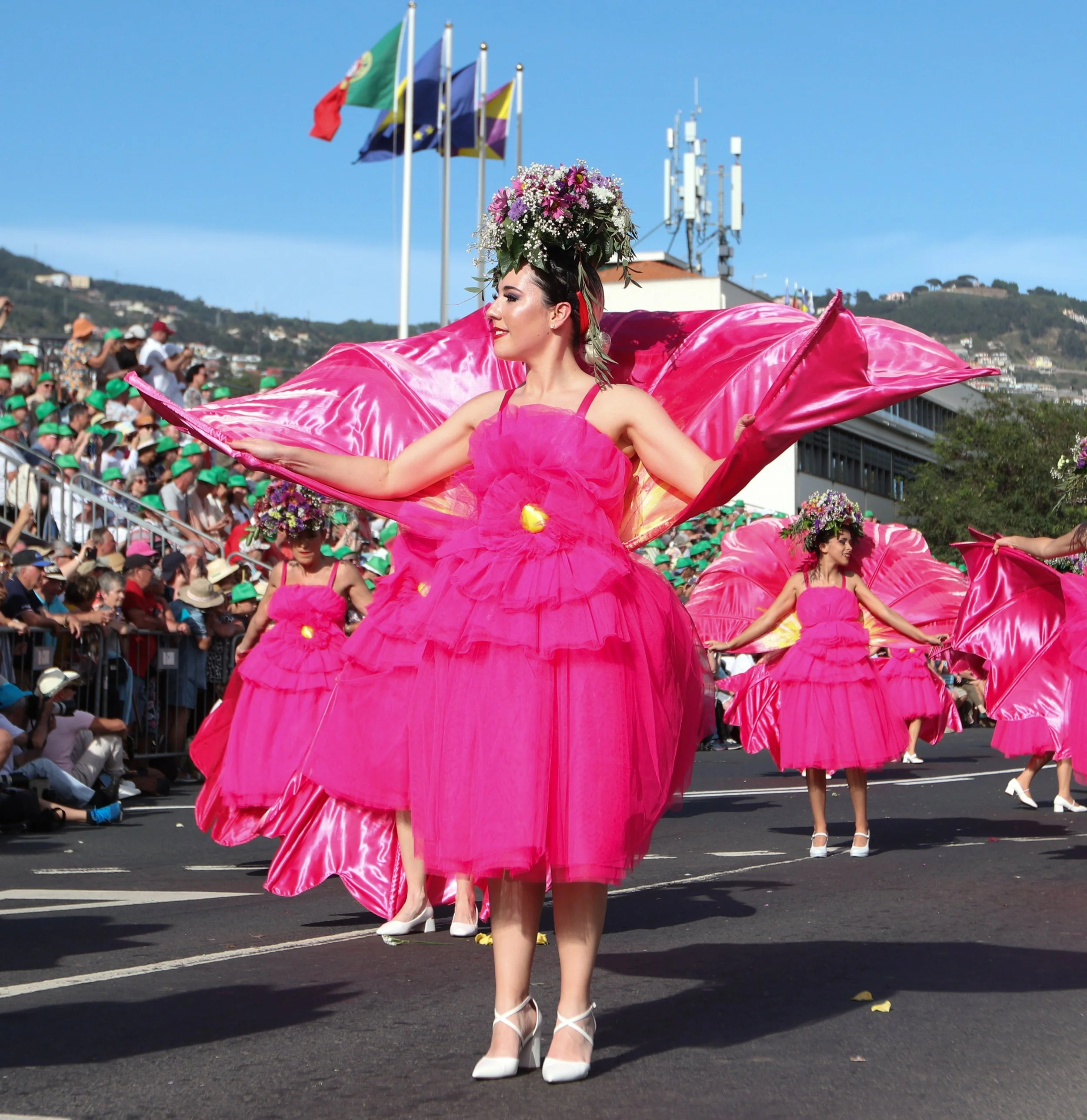 Women dressed in bright pink tutus and flower crowns performing a parade dance on a city street, with a crowd of spectators in the background and flags flying.